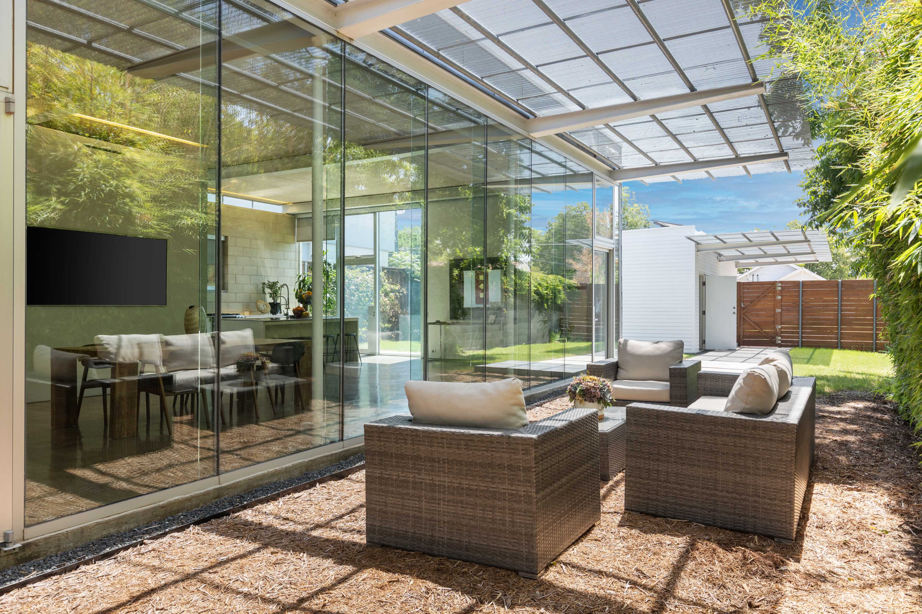 The image shows a modern outdoor living space with wicker furniture and a glass wall overlooking a green garden.