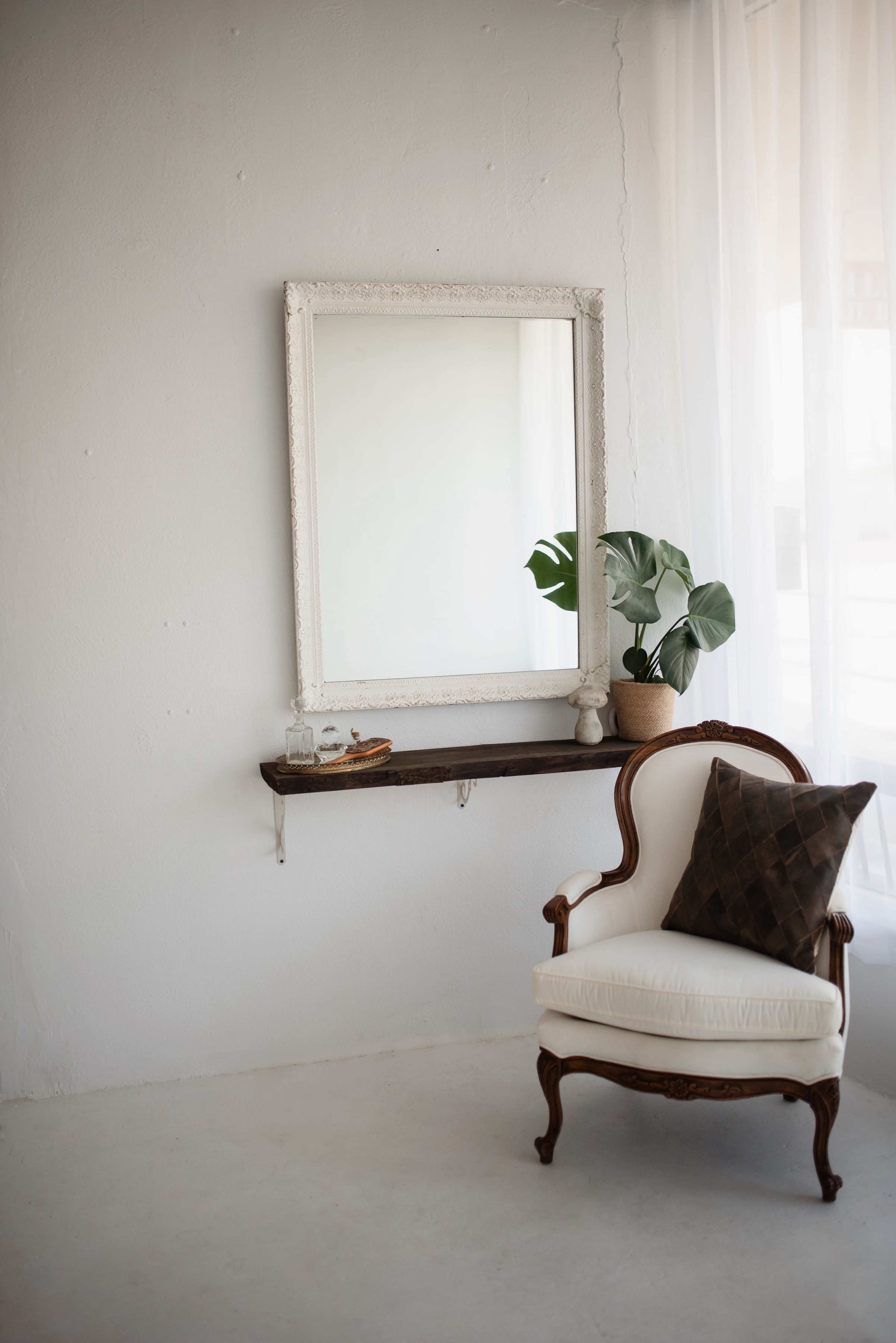 A vintage chair is positioned next to a wall-mounted mirror and a shelf adorned with a potted plant in a minimalist room.