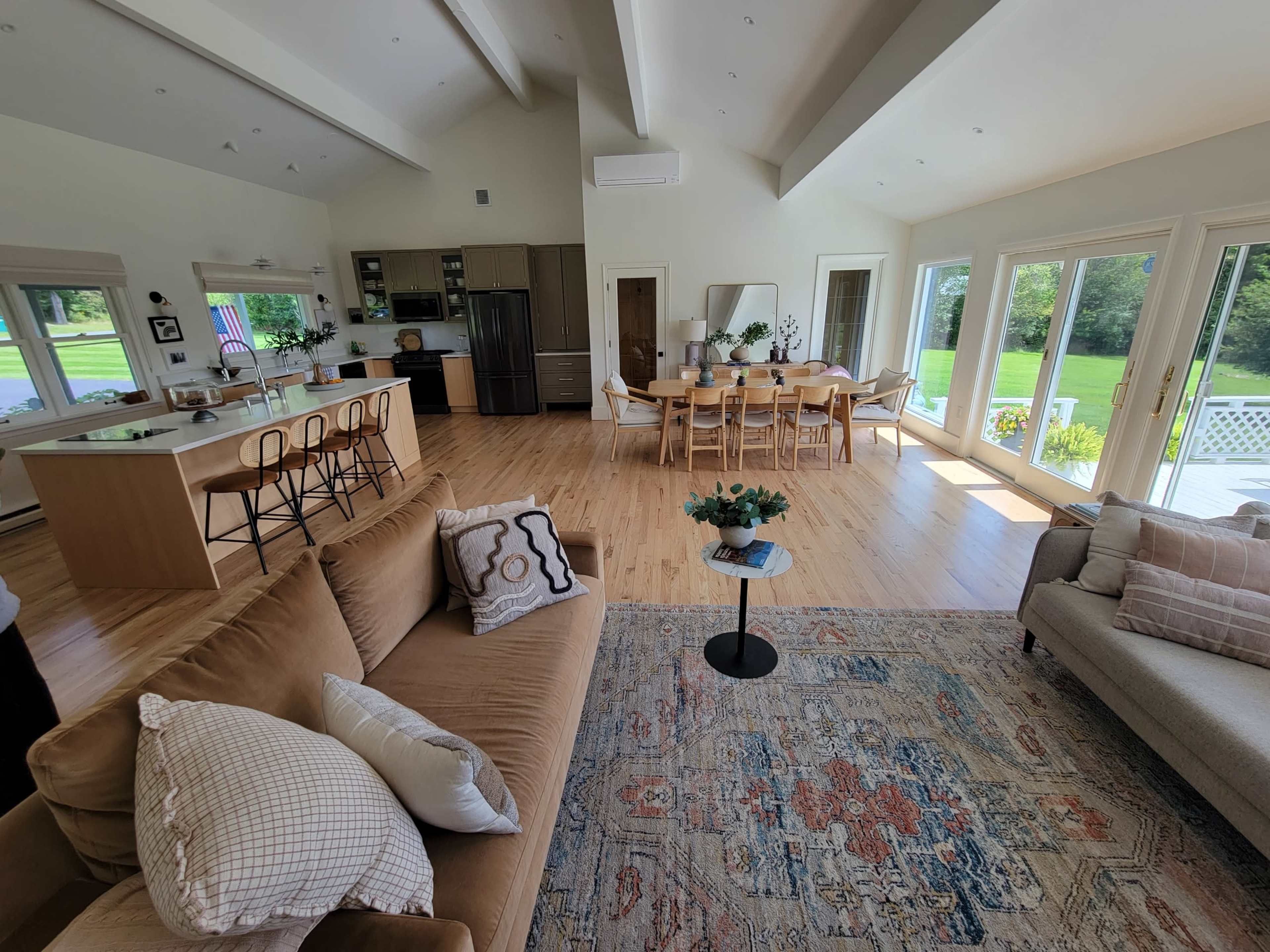 A spacious living area features a sofa, coffee table, and a dining table with chairs, adjoining a kitchen with modern appliances and large windows overlooking a garden.