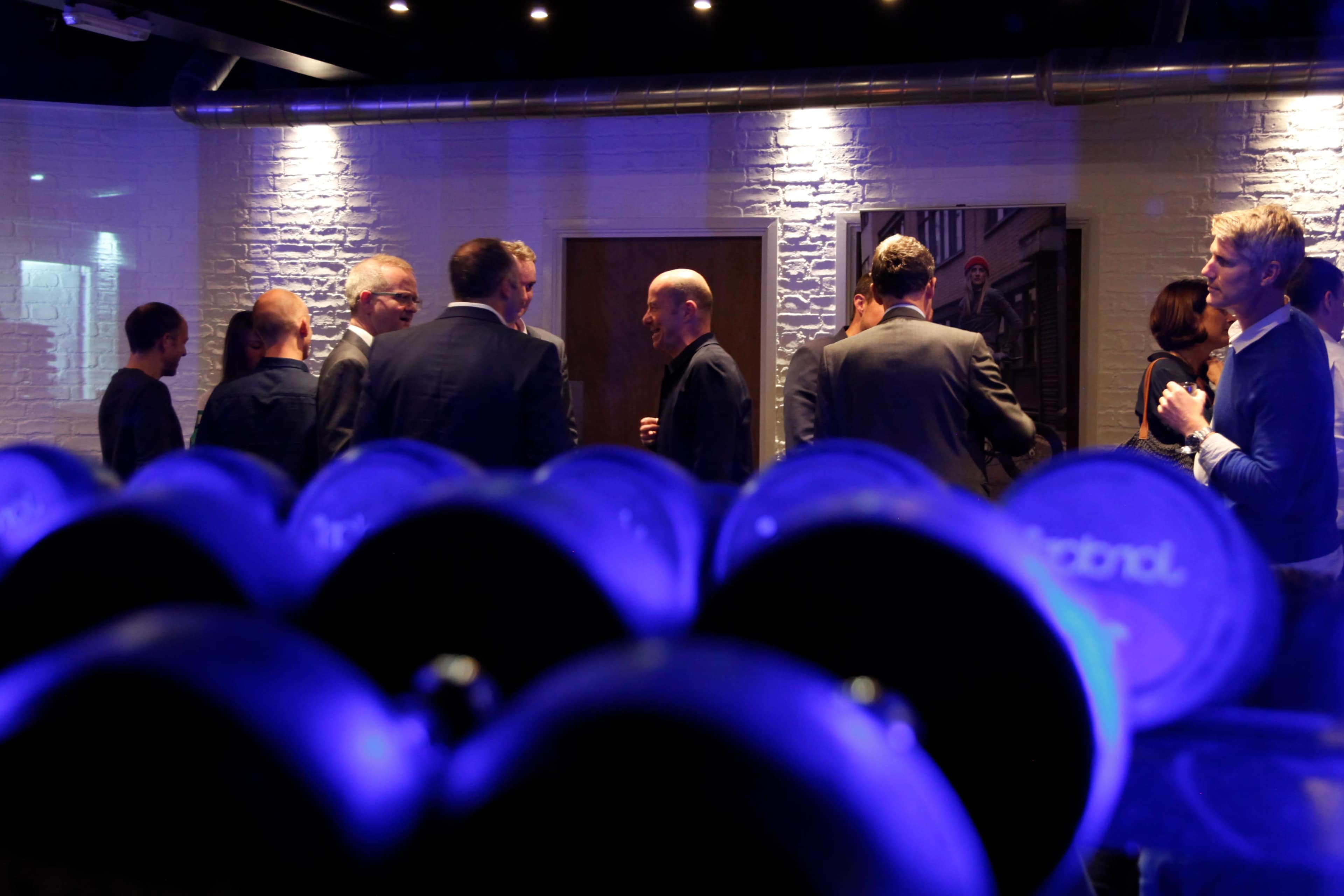 A group of people engage in conversation in a dimly lit room, with weights in the foreground.