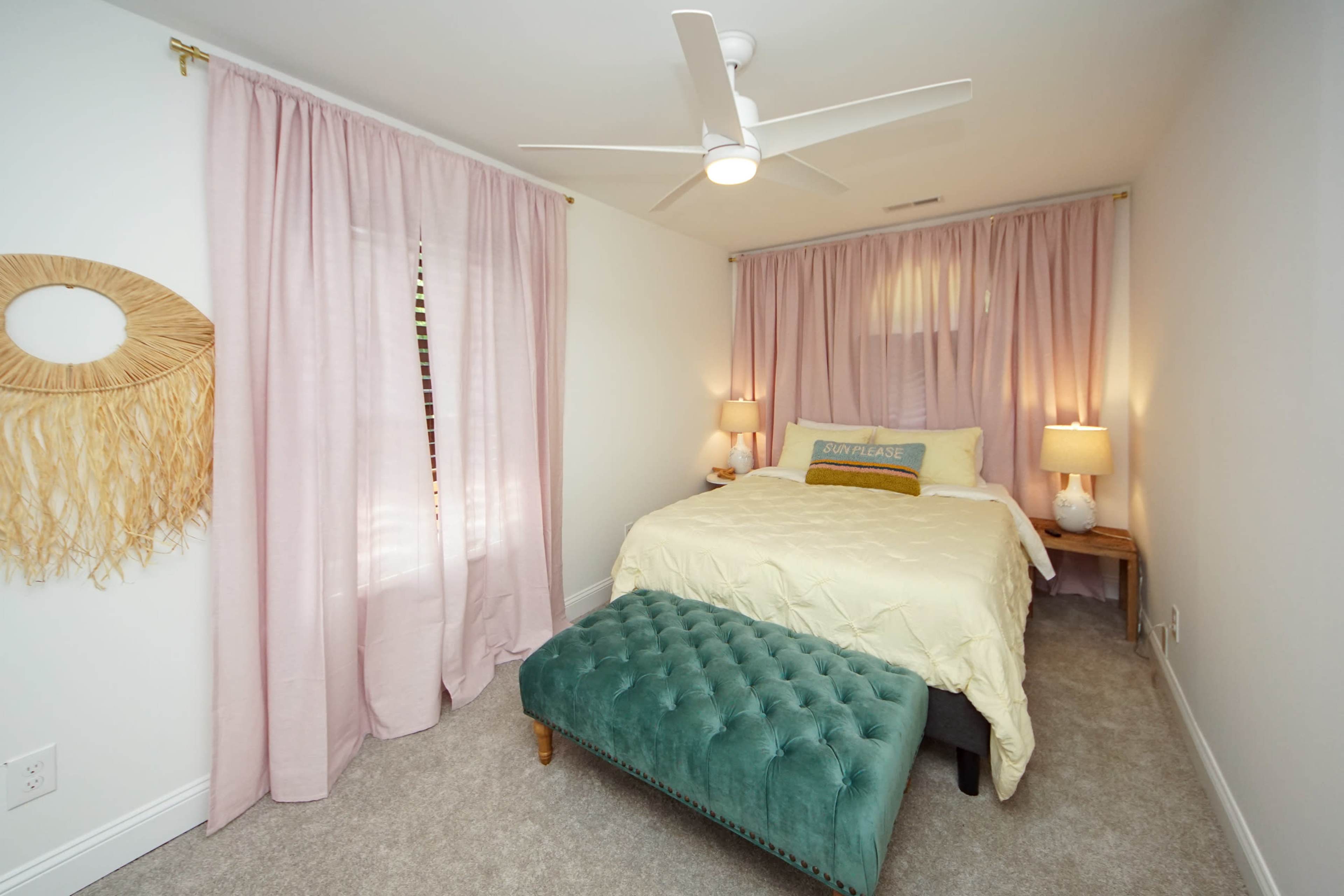 A cozy bedroom featuring a bed with a light yellow comforter, pink curtains, a green tufted bench, and two bedside lamps.