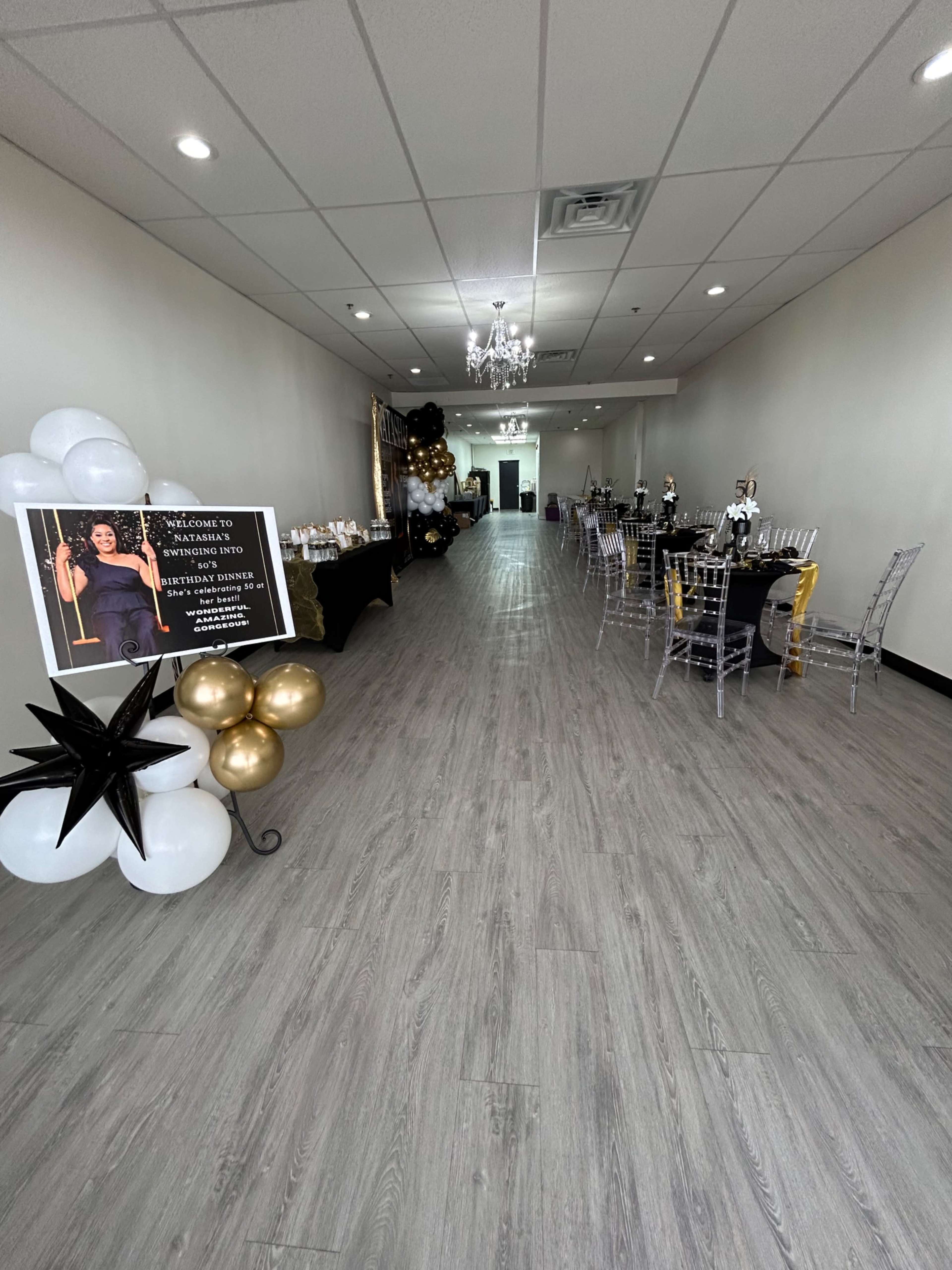 A long, narrow event space decorated with black and gold balloons, a sign welcoming guests, and tables set for an event along the right side.