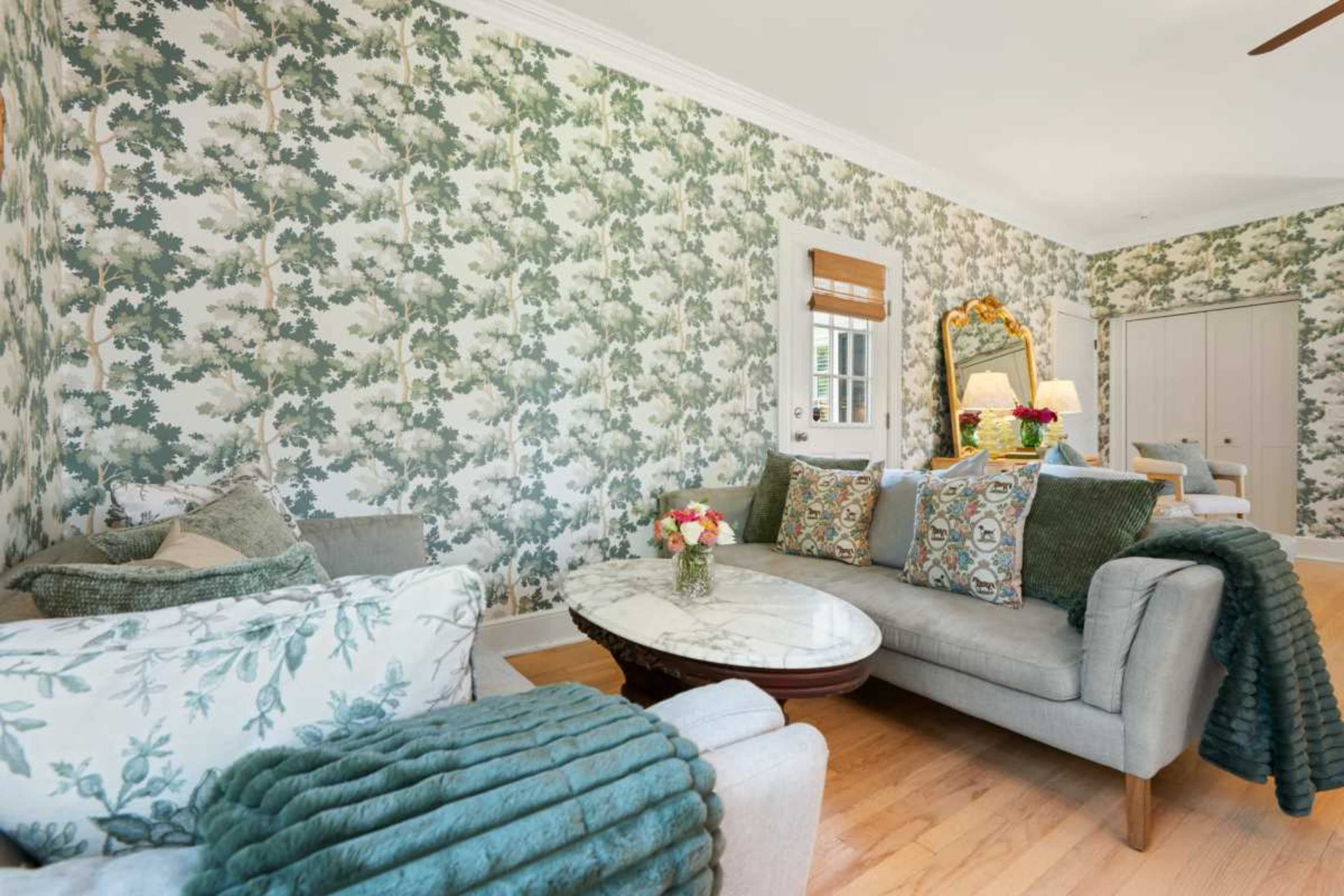 A living room features gray sofas adorned with decorative pillows, a marble coffee table, and walls covered in green leafy wallpaper.