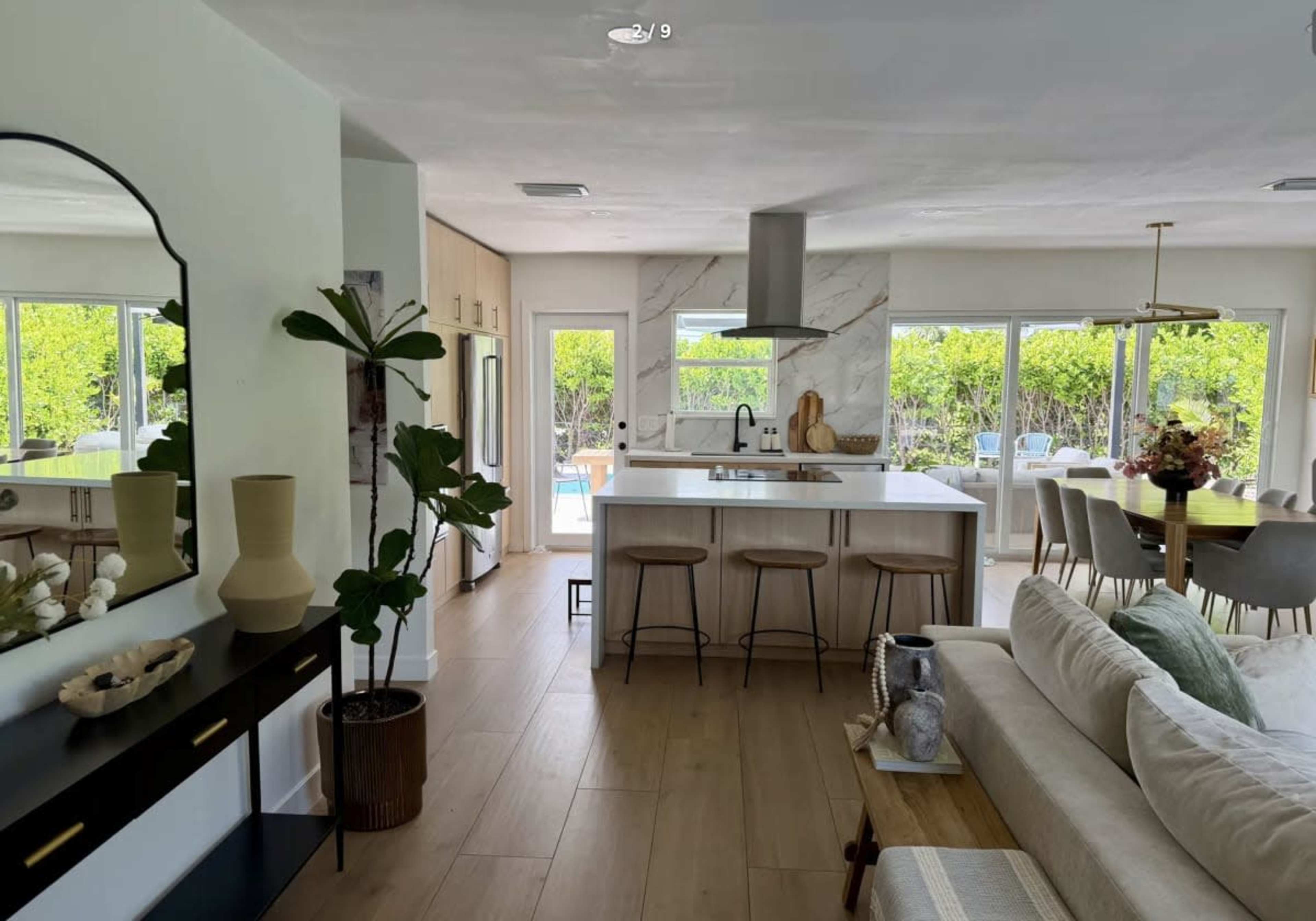 The image shows a modern open-concept kitchen and living area, featuring a large island with stools, a dining table in the background, and sliding glass doors leading to an outdoor space.