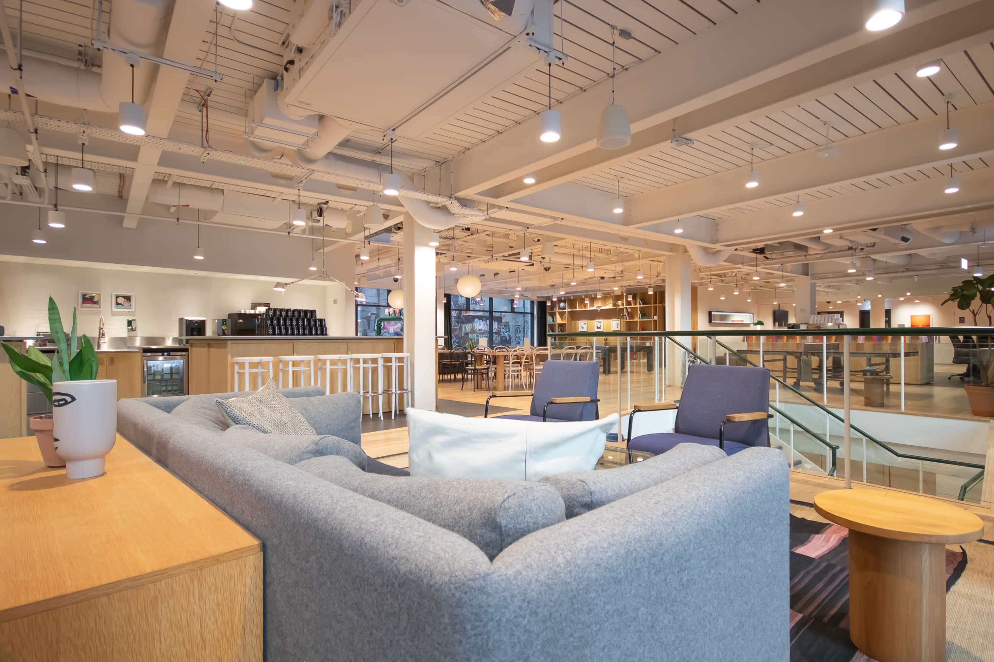 The image depicts a modern, open-plan office space with comfortable seating areas, wooden tables, and a cafe area in the background.