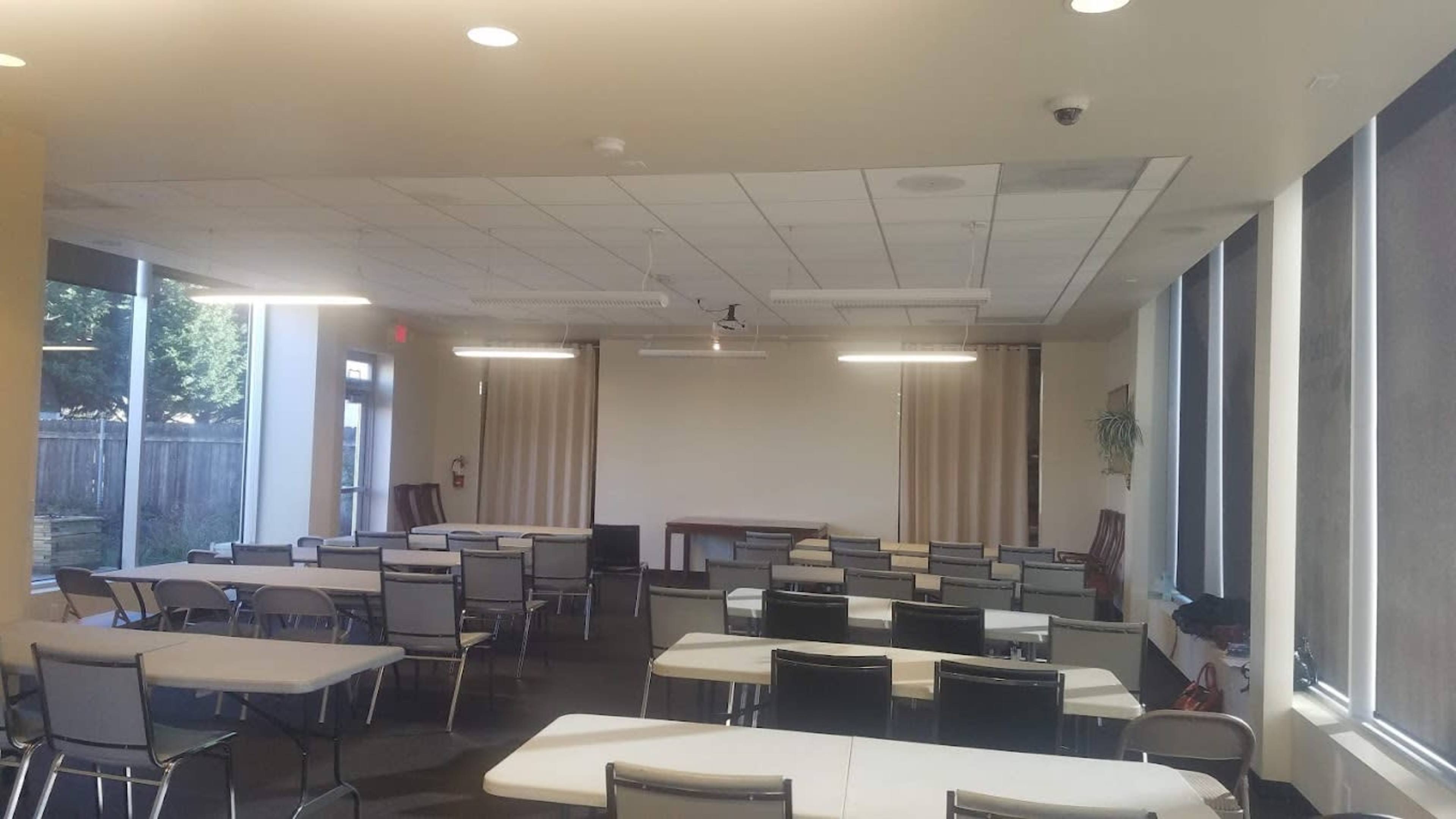 A conference room with rows of tables and chairs arranged for a meeting, featuring large windows and a blank wall at the front.