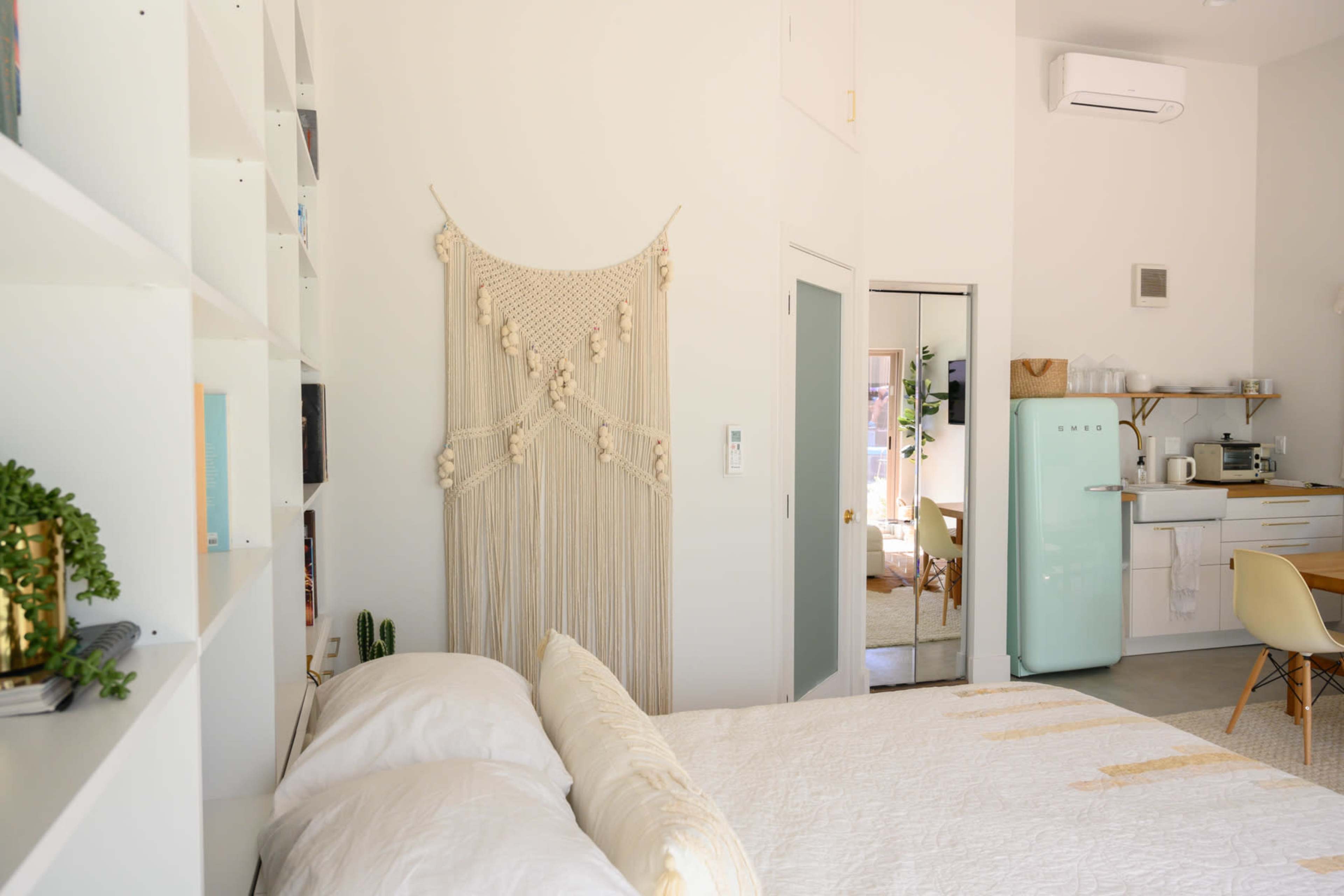 A bright, modern studio apartment features a bed with white linens, a macramé wall hanging, and a kitchenette with a retro-style refrigerator.