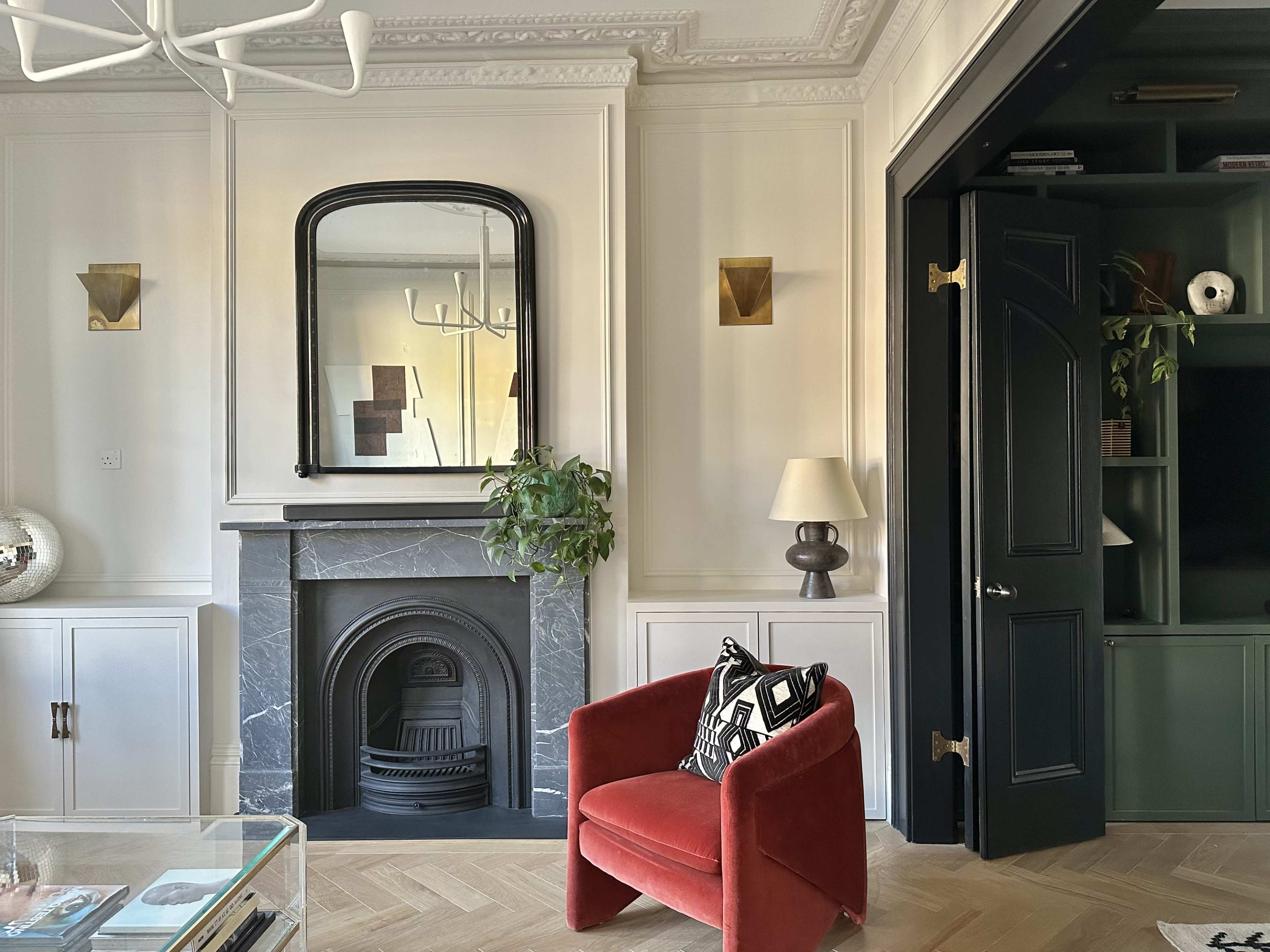 The image shows a stylish living room featuring a red armchair, a black marble fireplace with a mirror above it, and dark green shelving adjacent to a black door.