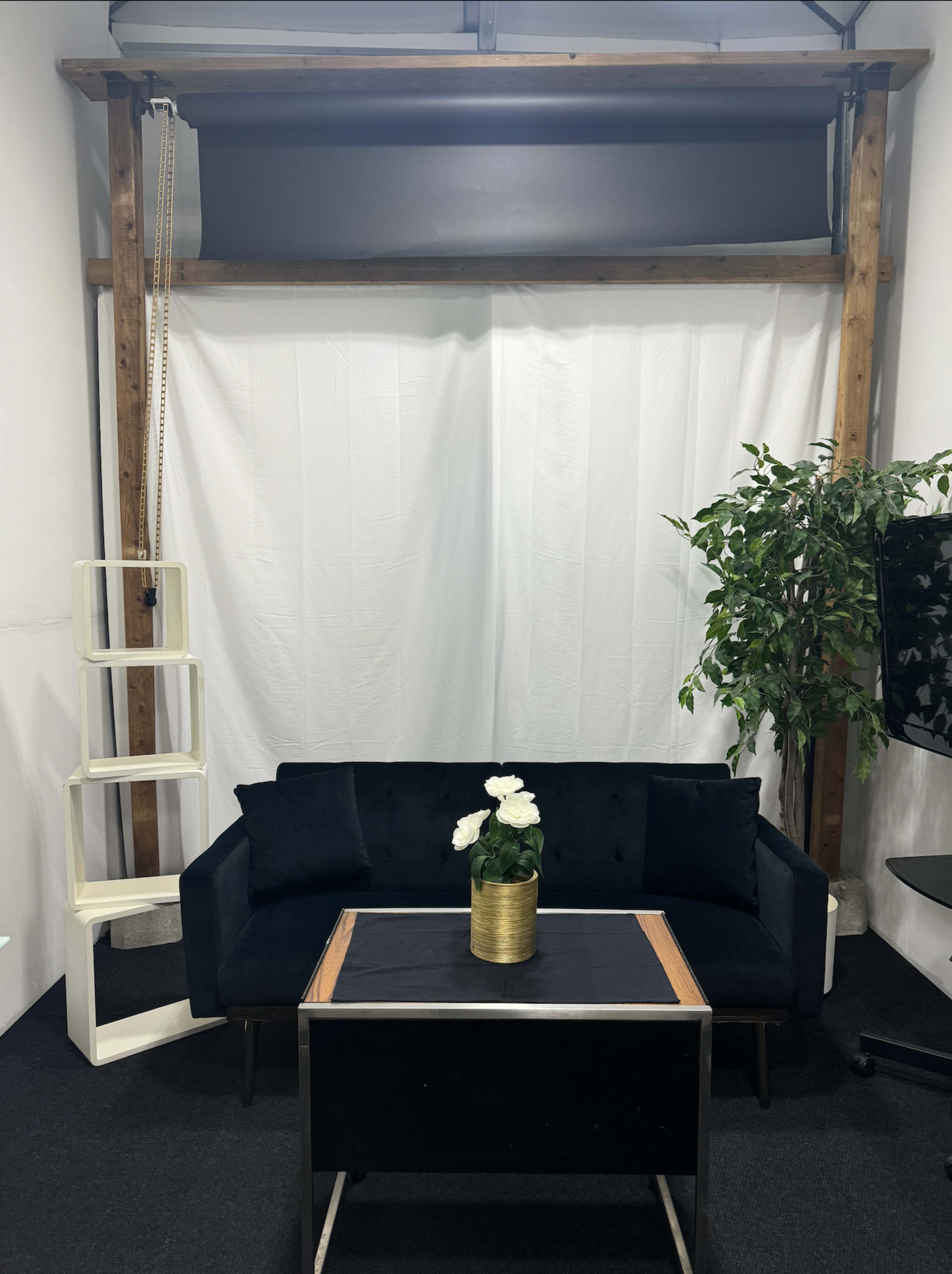 The image shows a minimalist interior featuring a black couch, a glass-top coffee table, a potted plant, and a white backdrop with wooden framing.