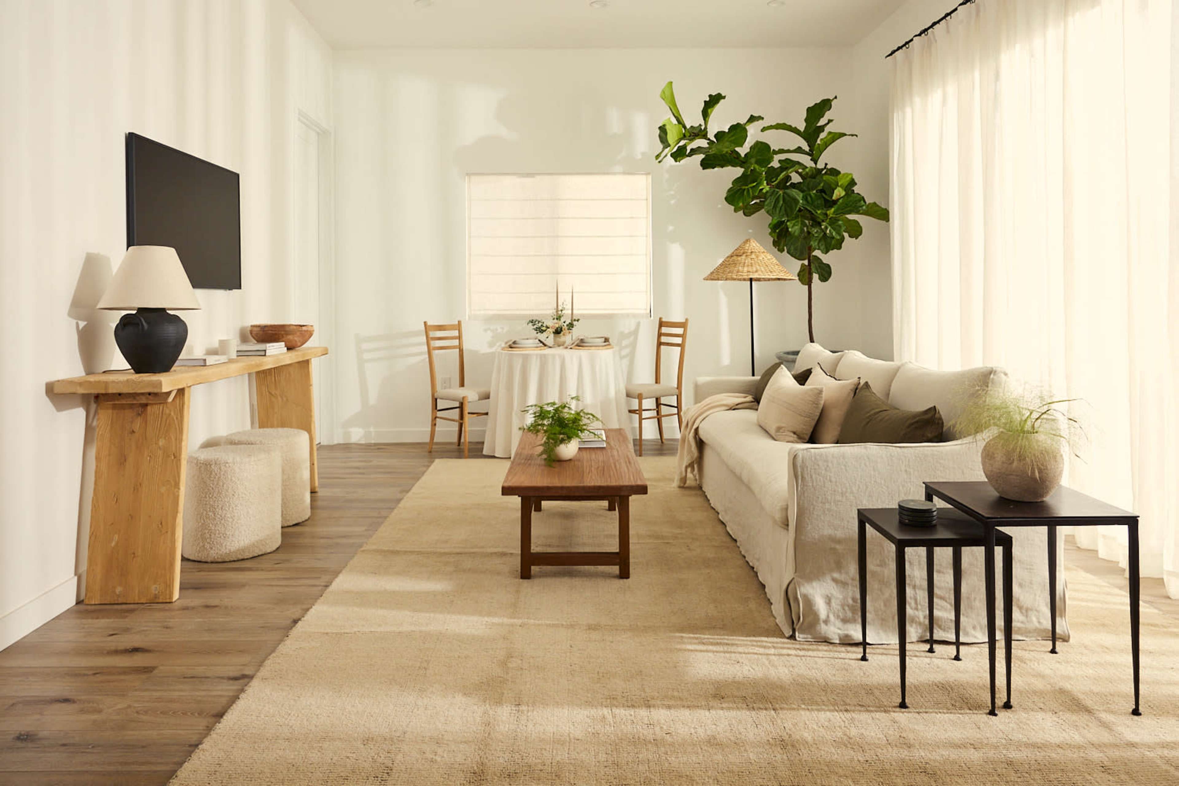 A light-filled living room features a neutral color palette with a sofa, coffee table, two side tables, and a dining area in the background.