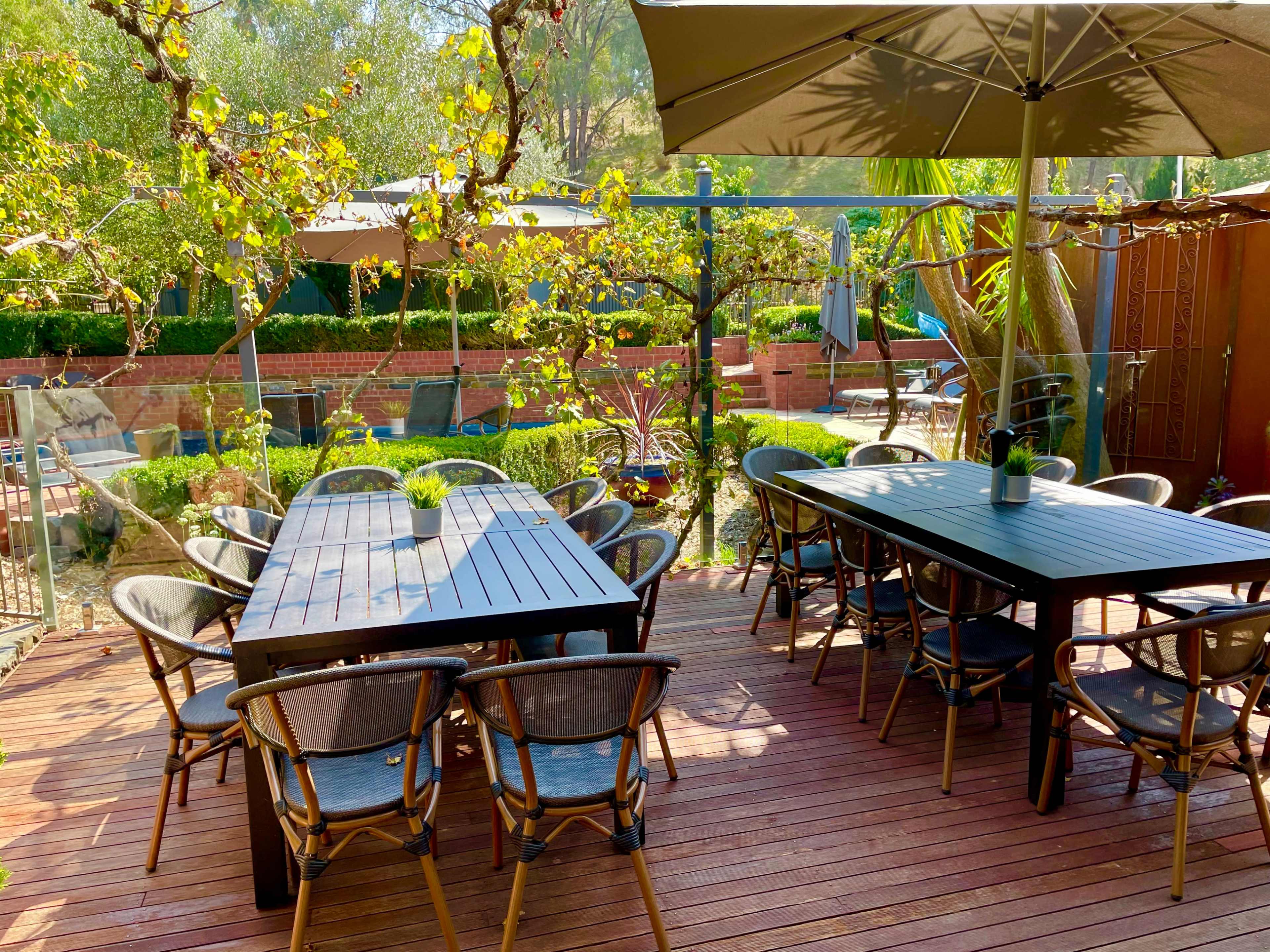 The outdoor dining area features several black wooden tables surrounded by chairs, set against a backdrop of greenery and a pool area.