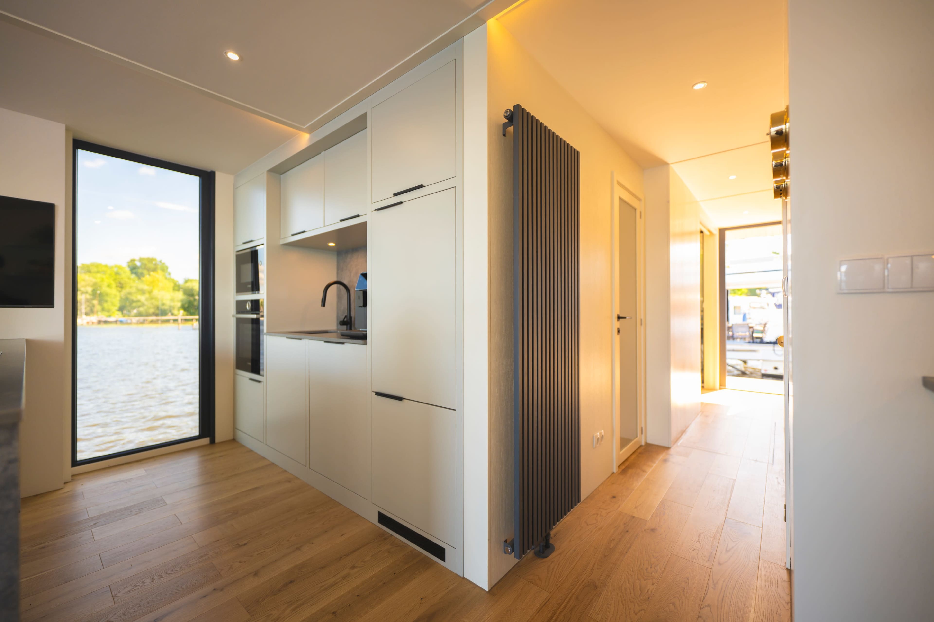 The image shows a modern kitchen with sleek cabinetry, positioned adjacent to a hallway leading to an outdoor view by the water.