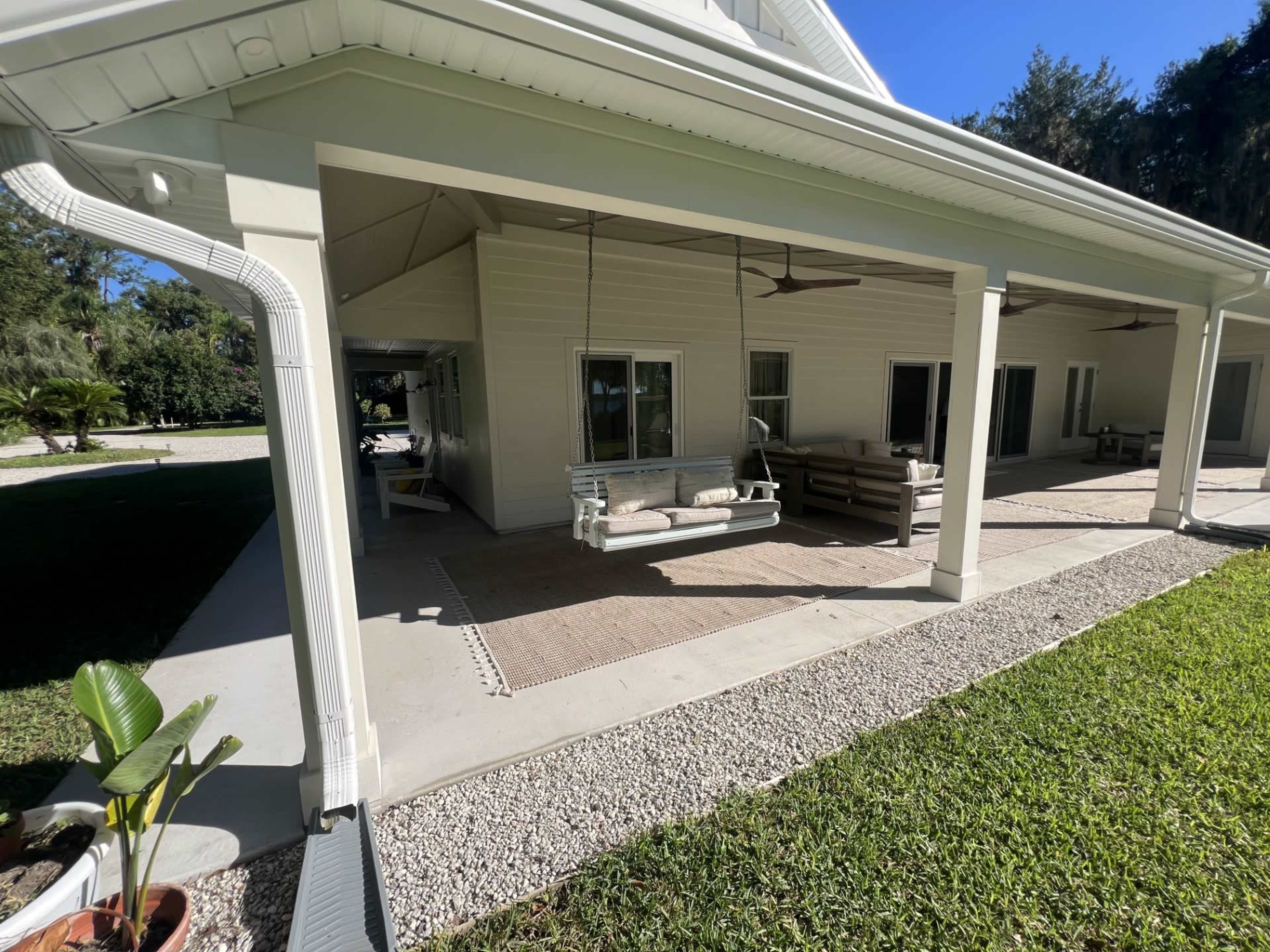 The image shows a spacious porch area of a house with a swing, seating, and decorative elements, surrounded by a lawn and trees.