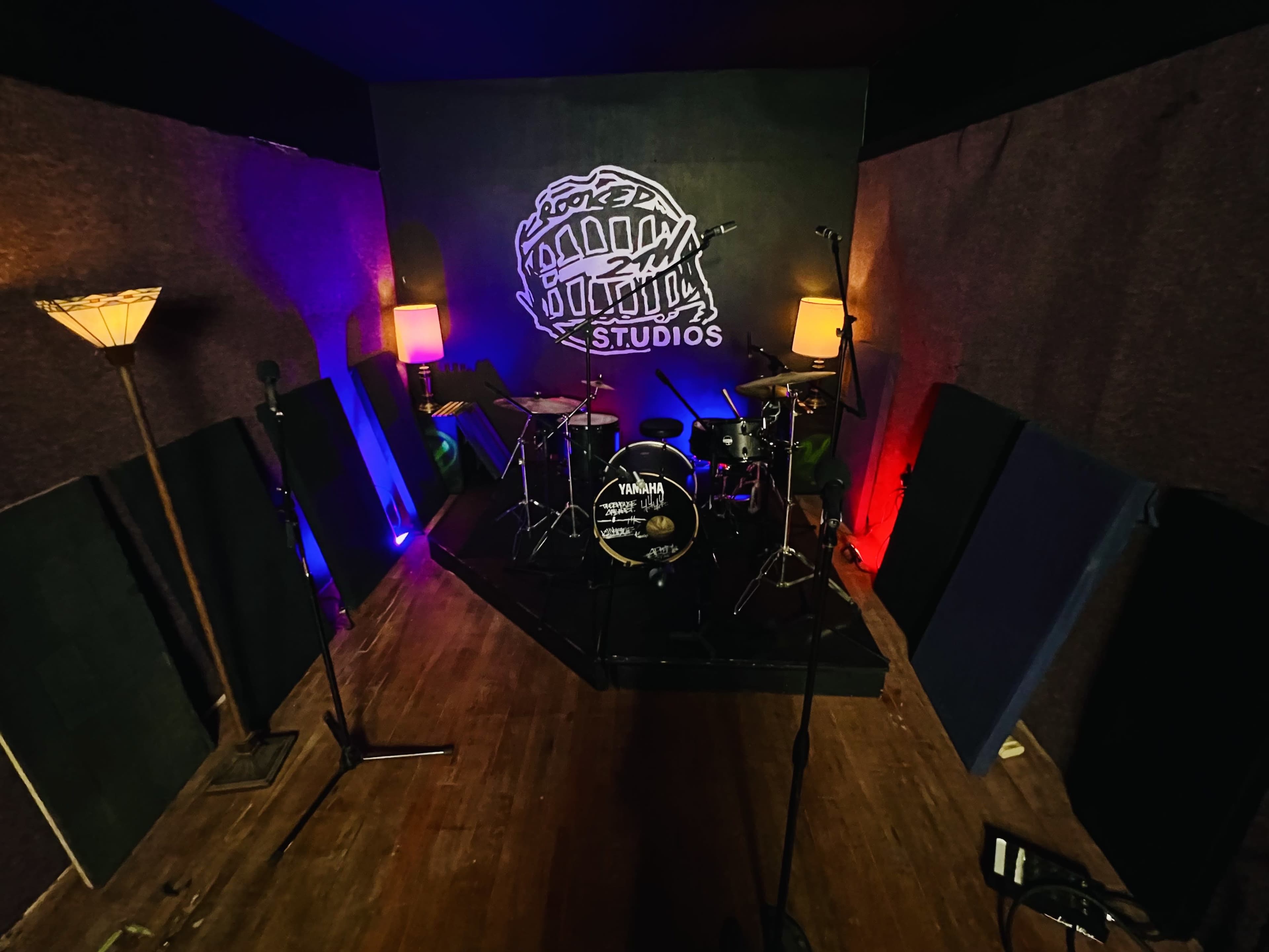 The image shows a small recording studio with a drum set, microphones, and soundproofing panels illuminated by lamplight.
