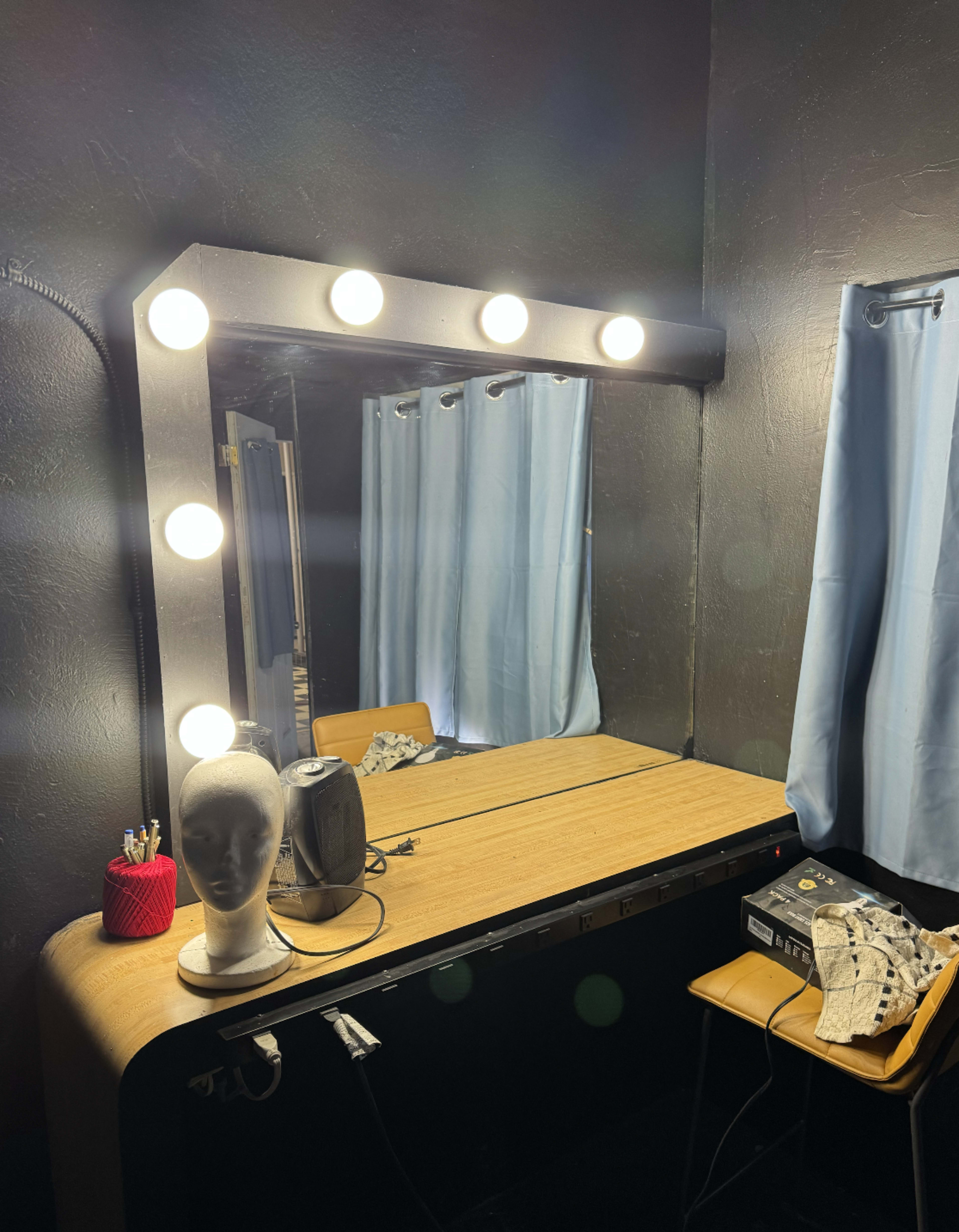 The image shows a well-lit vanity mirror with a wooden table, a mannequin head, and a few accessories on the surface, set against a dark wall and blue curtains.
