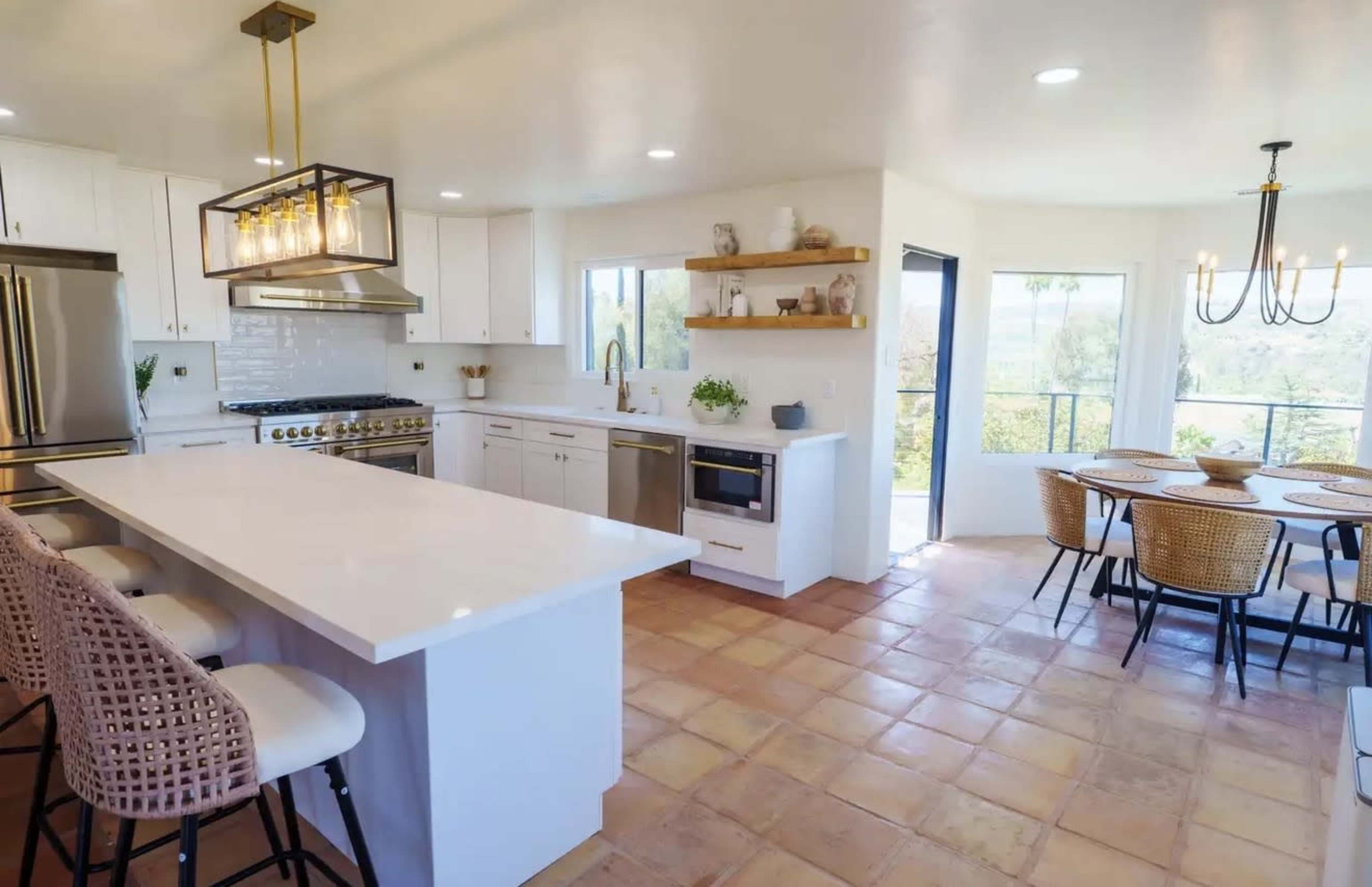 The image shows a modern kitchen with white cabinetry, a large island, stainless steel appliances, and a dining area with a round table and woven chairs.