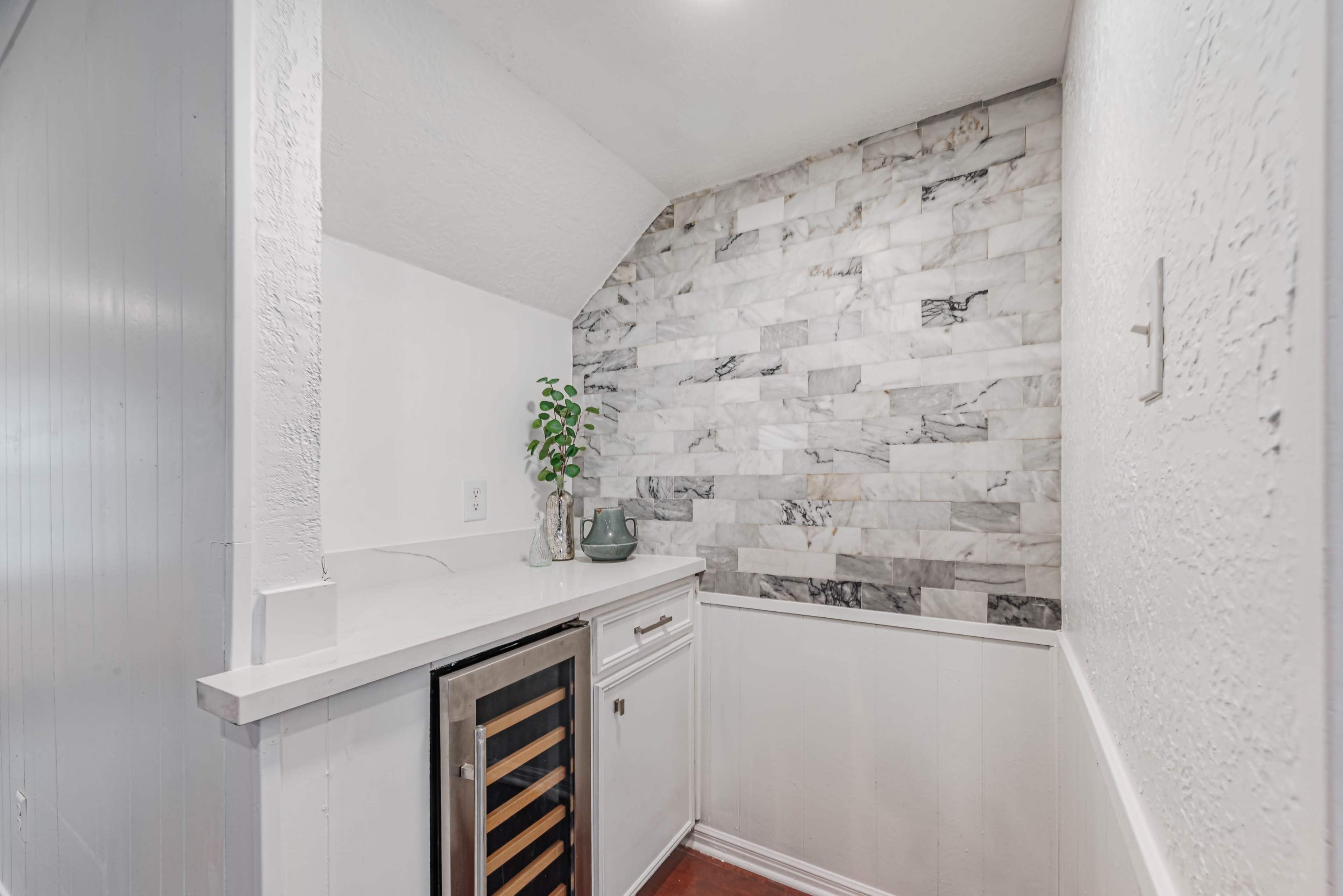 The image shows a small kitchen nook featuring a marble accent wall, a wine cooler, and minimal white cabinetry.