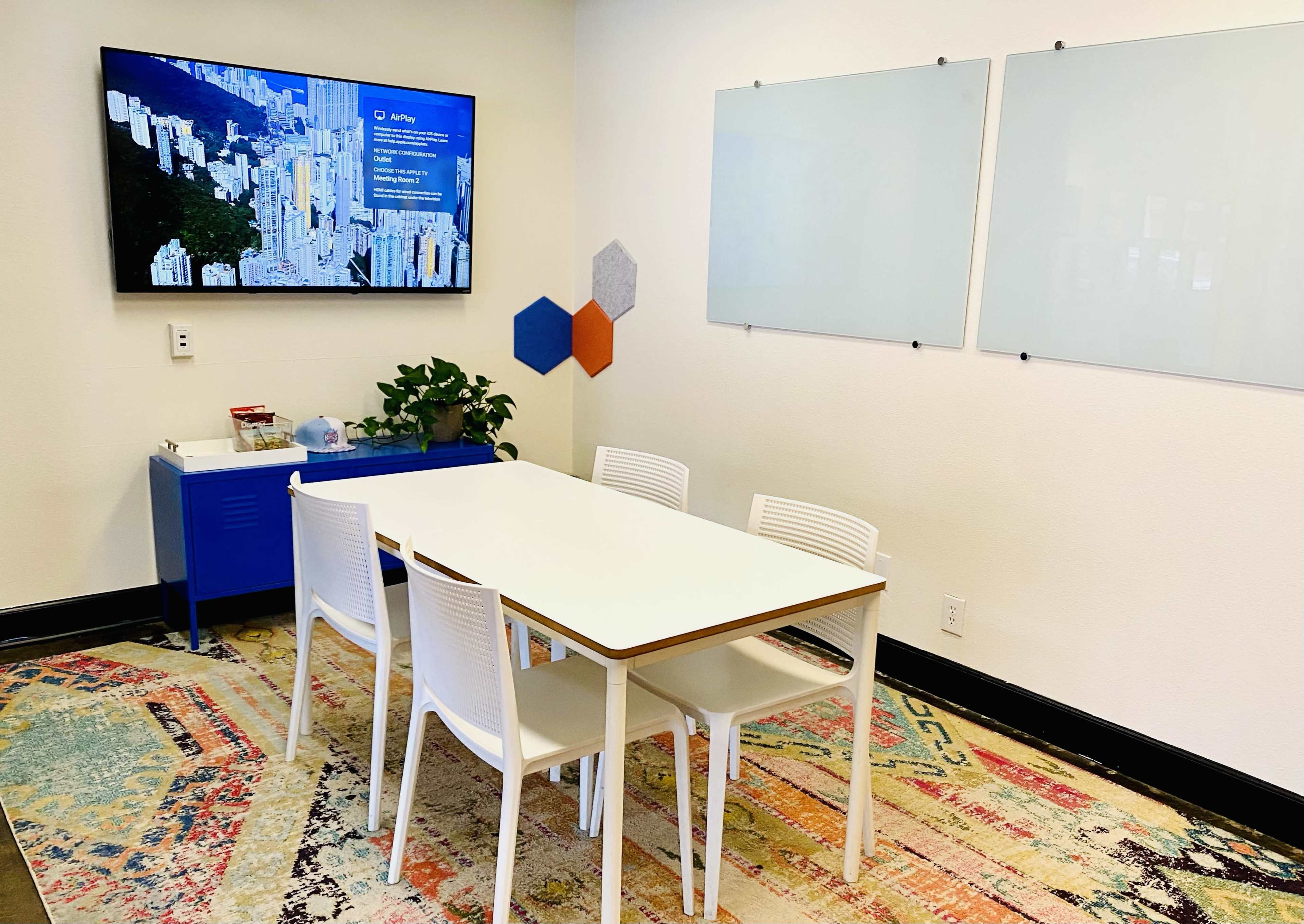 A conference room features a white table surrounded by four white chairs, a blue cabinet, and a television displaying a cityscape, with two empty whiteboards on the wall.