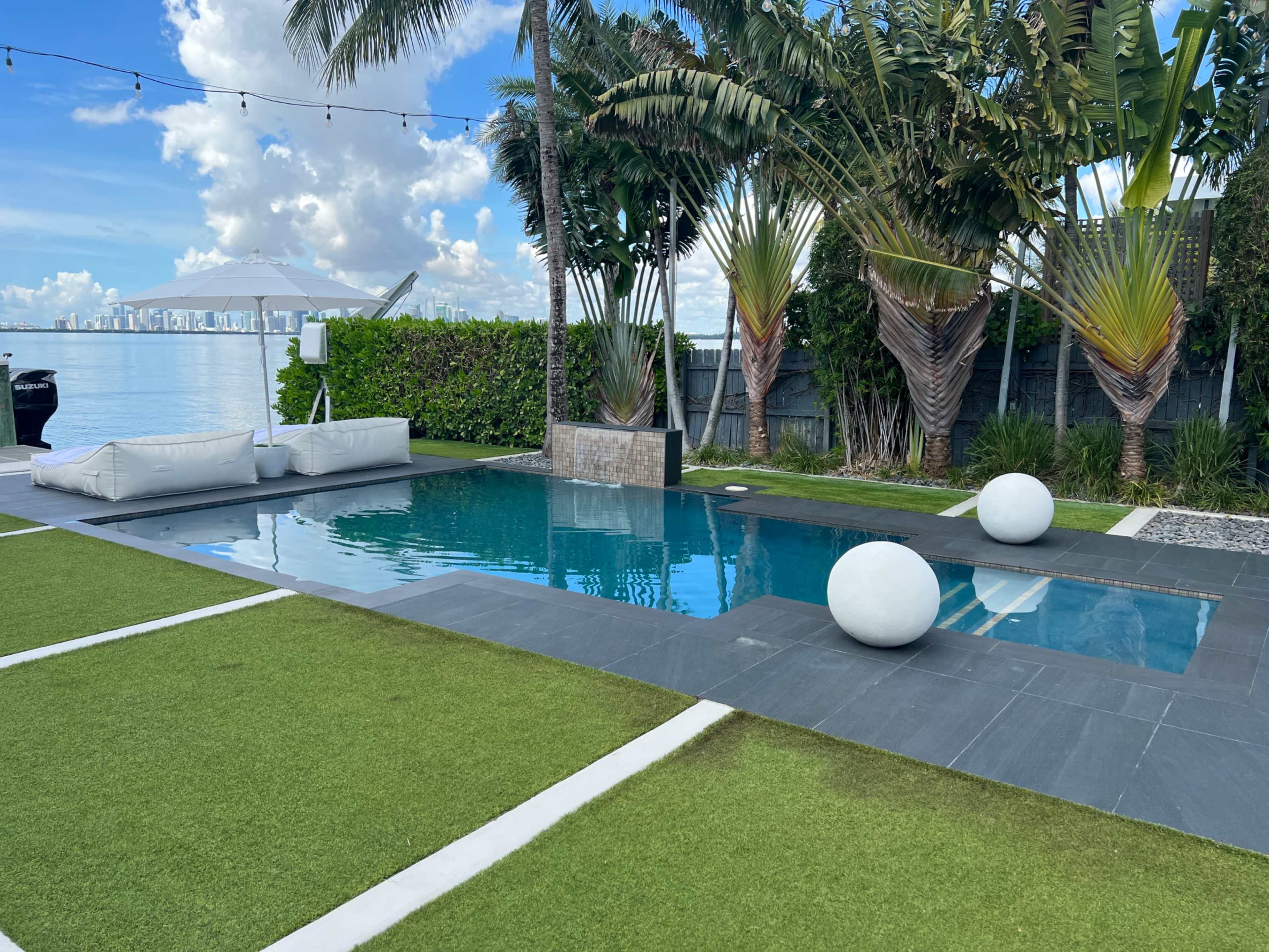 The image shows a modern pool area with a well-maintained lawn, two large white spherical decorations, and palm trees along the water's edge, with a city skyline visible in the background.