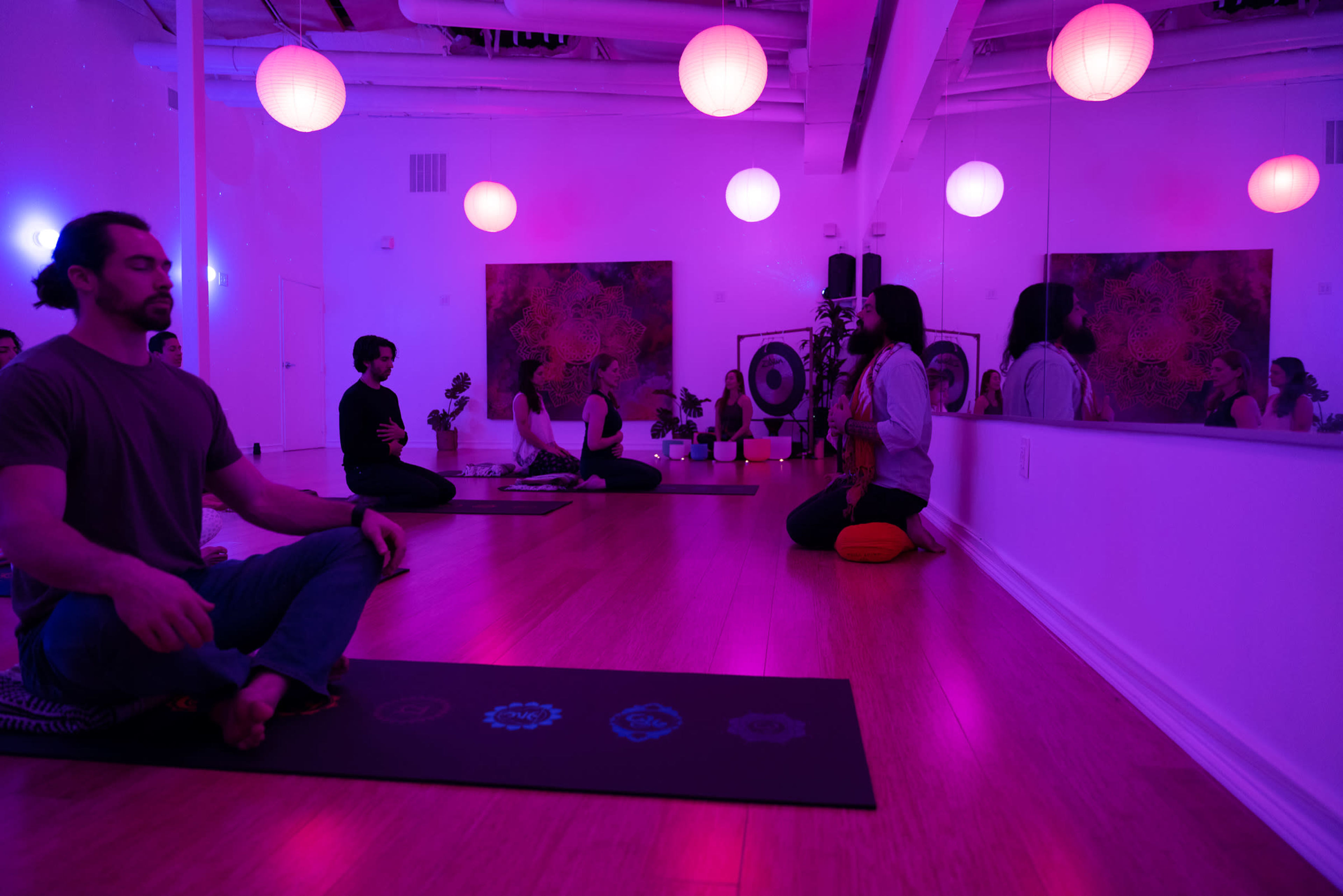 A group of individuals participates in a yoga or meditation session in a dimly lit studio adorned with colorful hanging lights.
