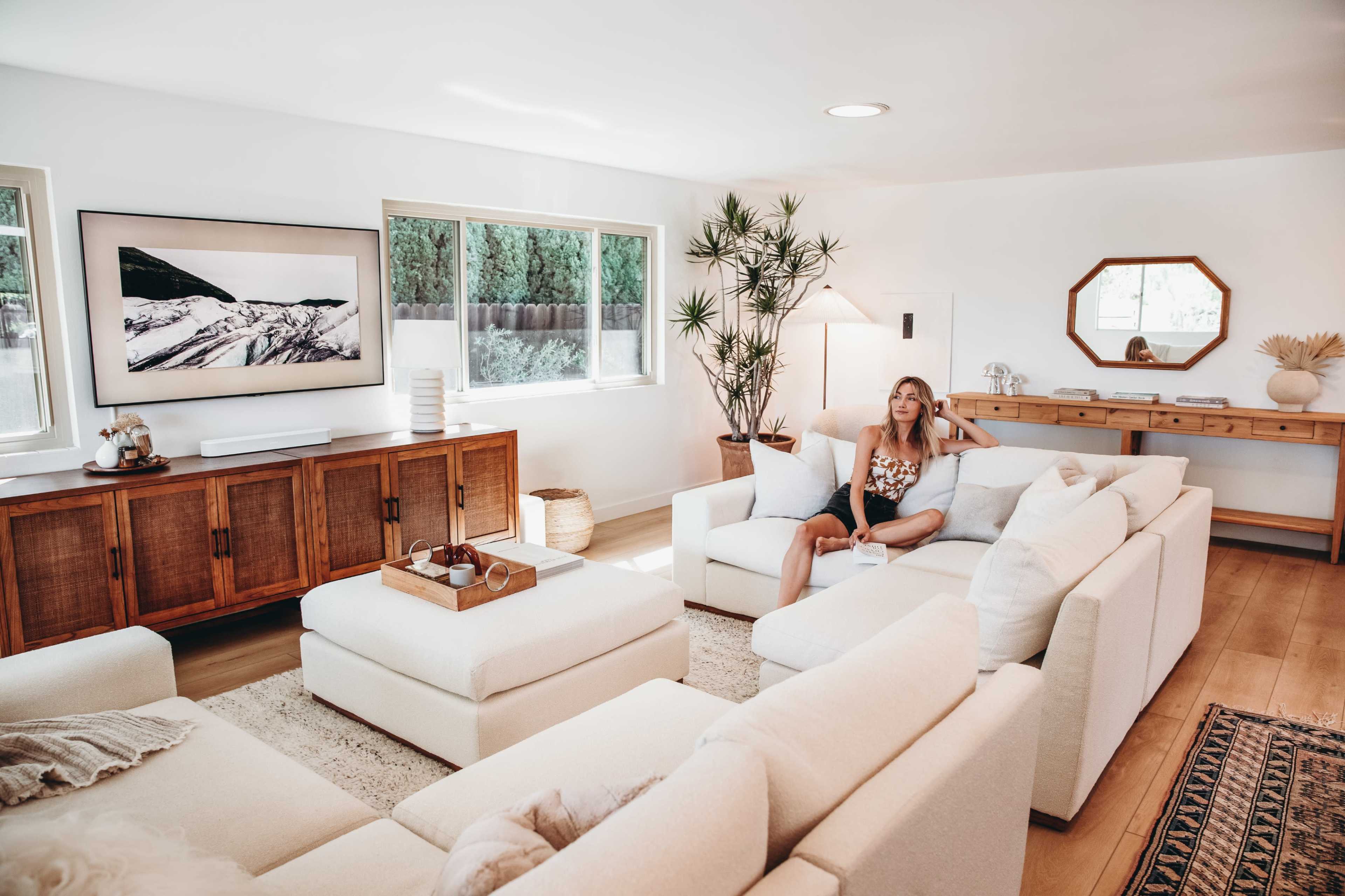 A woman sits on a light-colored sectional sofa in a bright living room with large windows and wooden furniture.