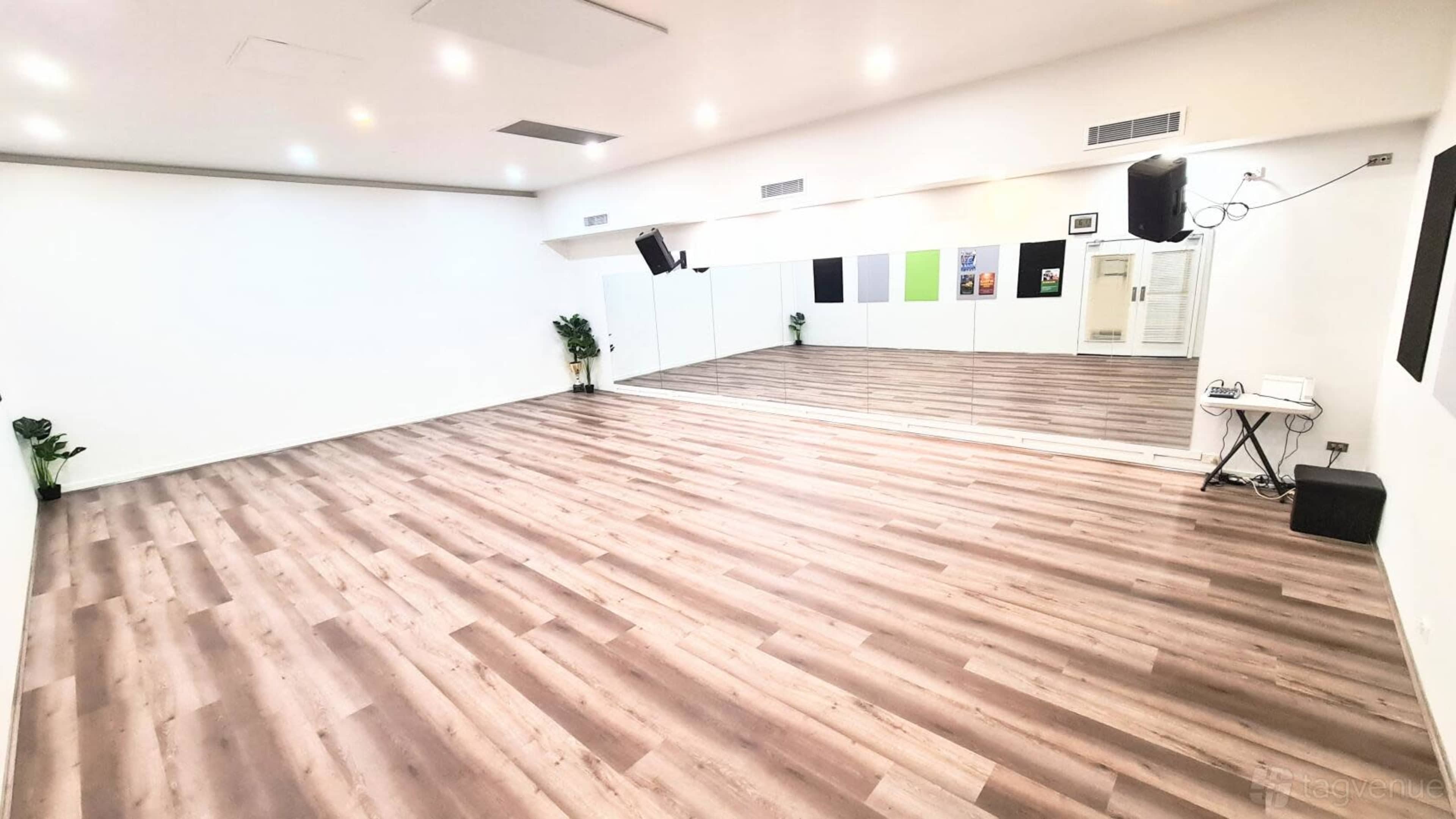 The image shows a spacious, empty dance studio with wooden flooring, mirrored walls, and minimal decor.