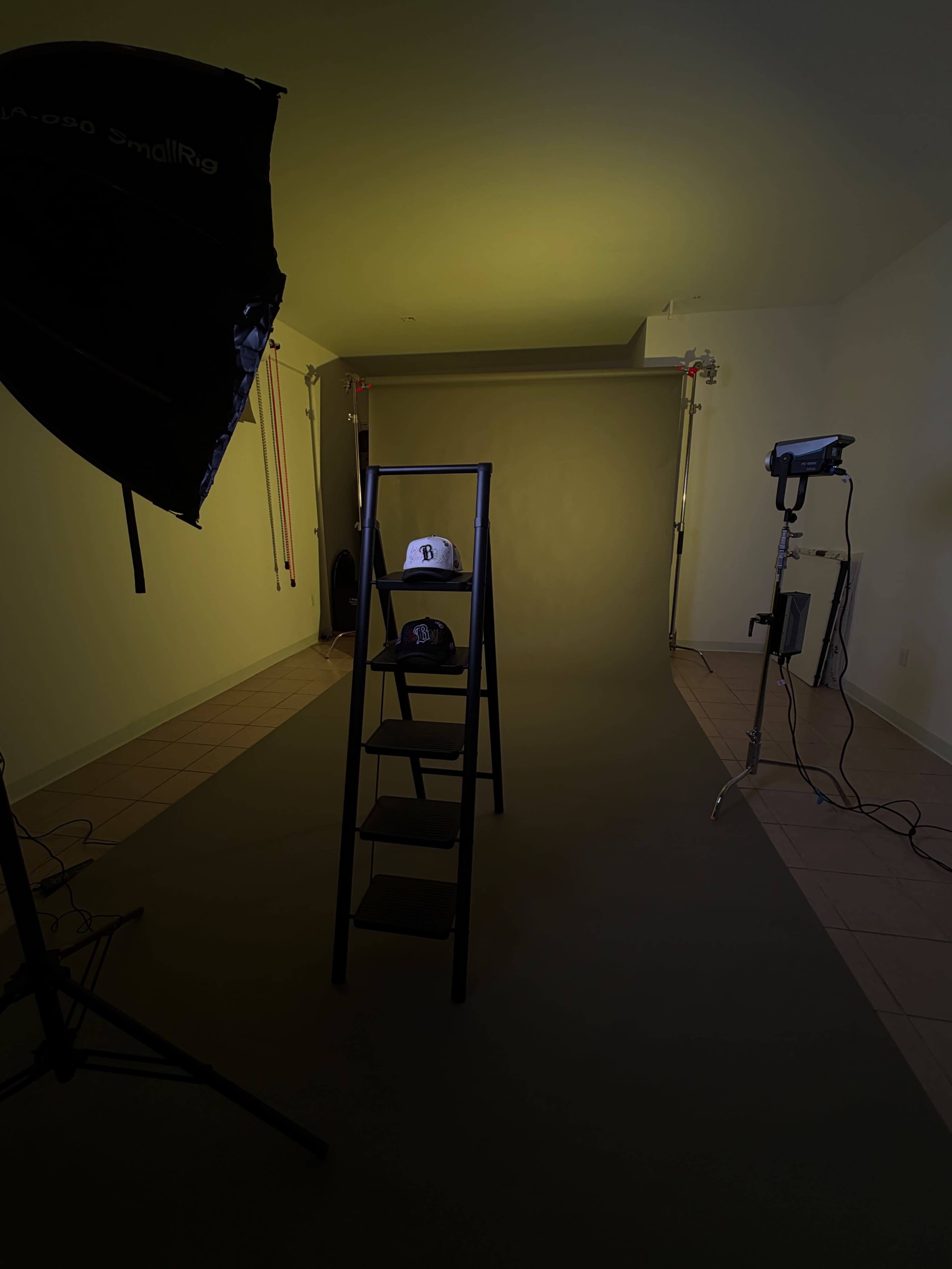A black ladder stands in front of a plain backdrop, displaying two hats under studio lights in a photography setup.