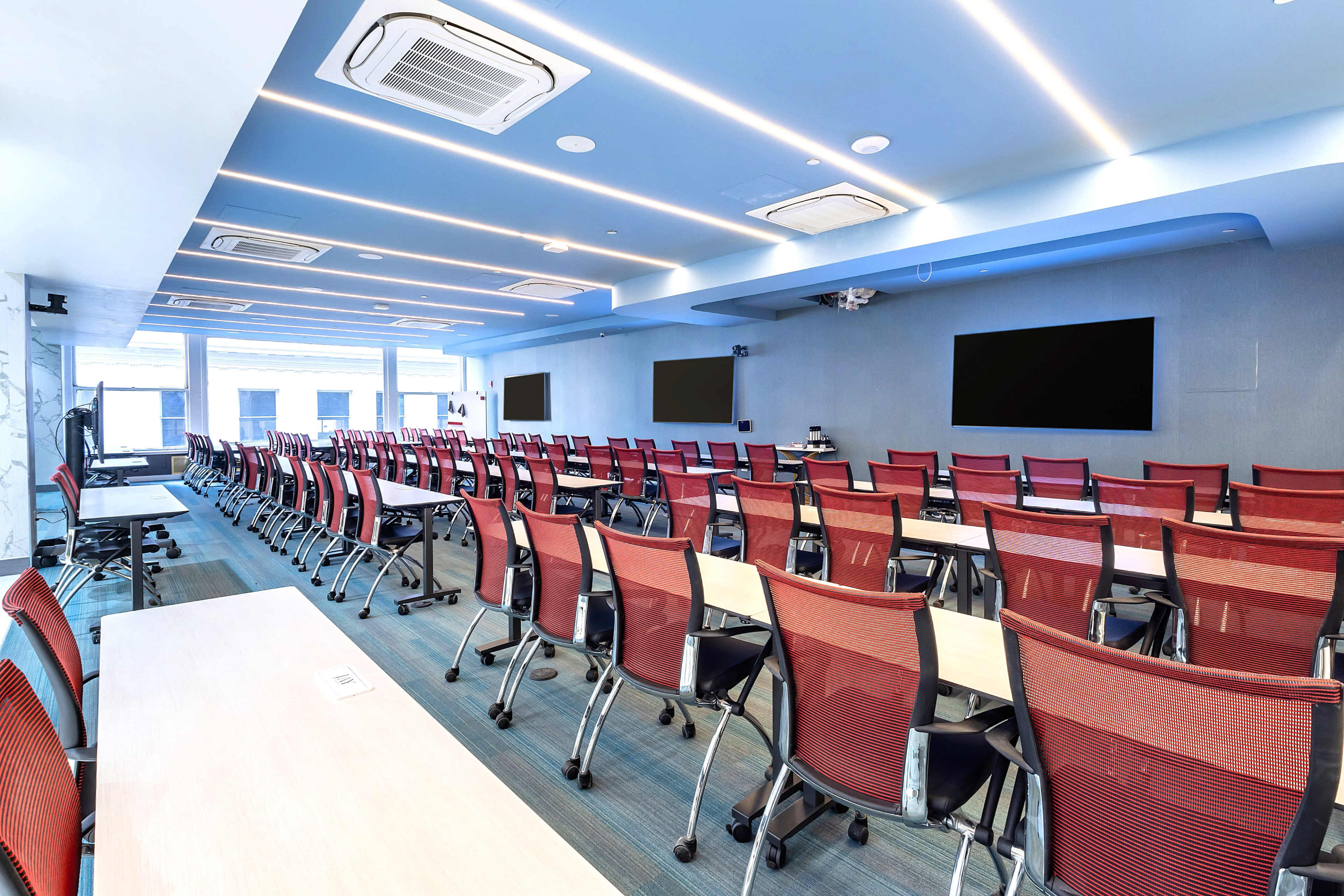 A modern conference room is set up with rows of red mesh chairs facing two large screens, under blue ambient lighting.