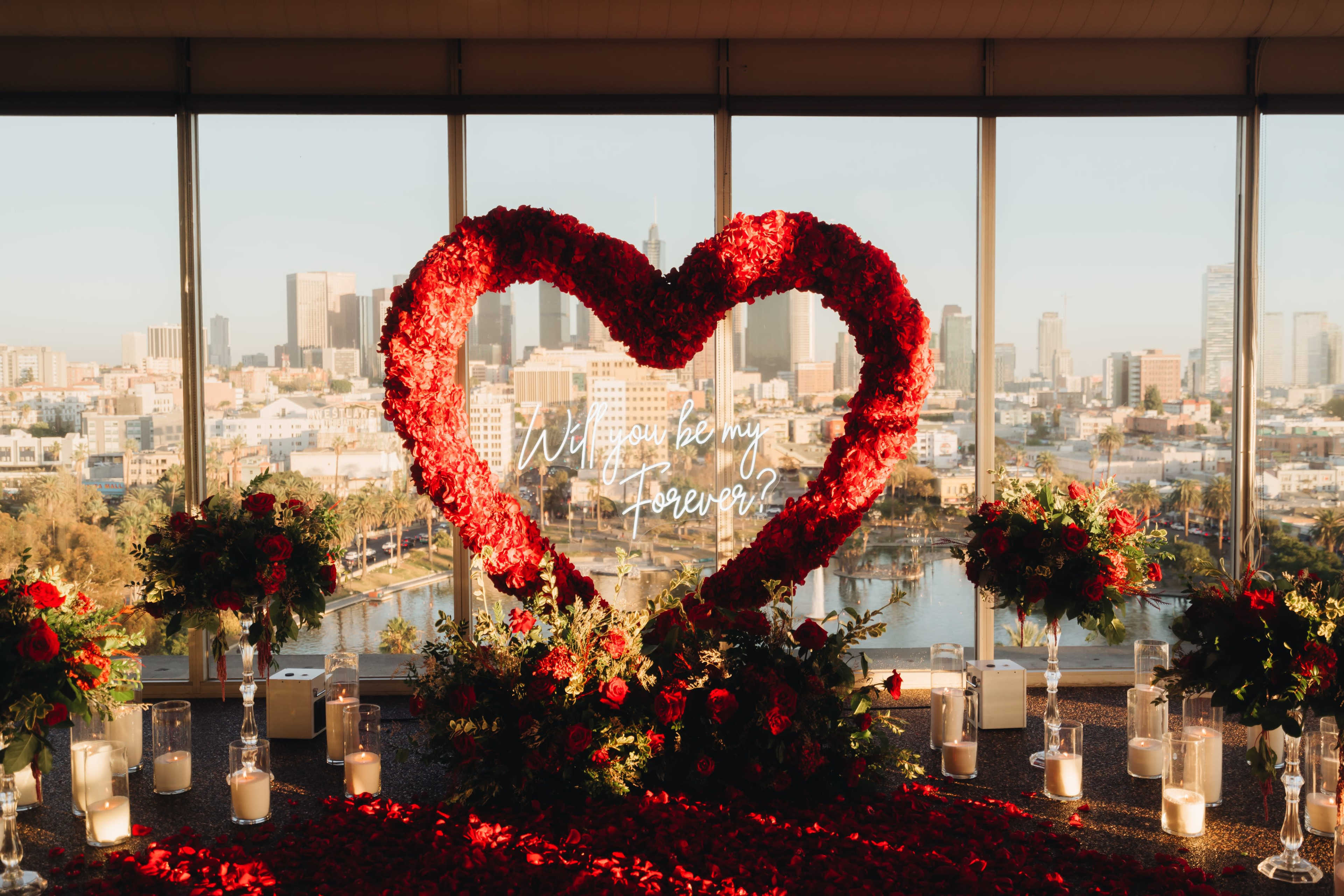 A heart-shaped floral arrangement adorned with red flowers and candles is displayed in front of large windows showcasing a city skyline.