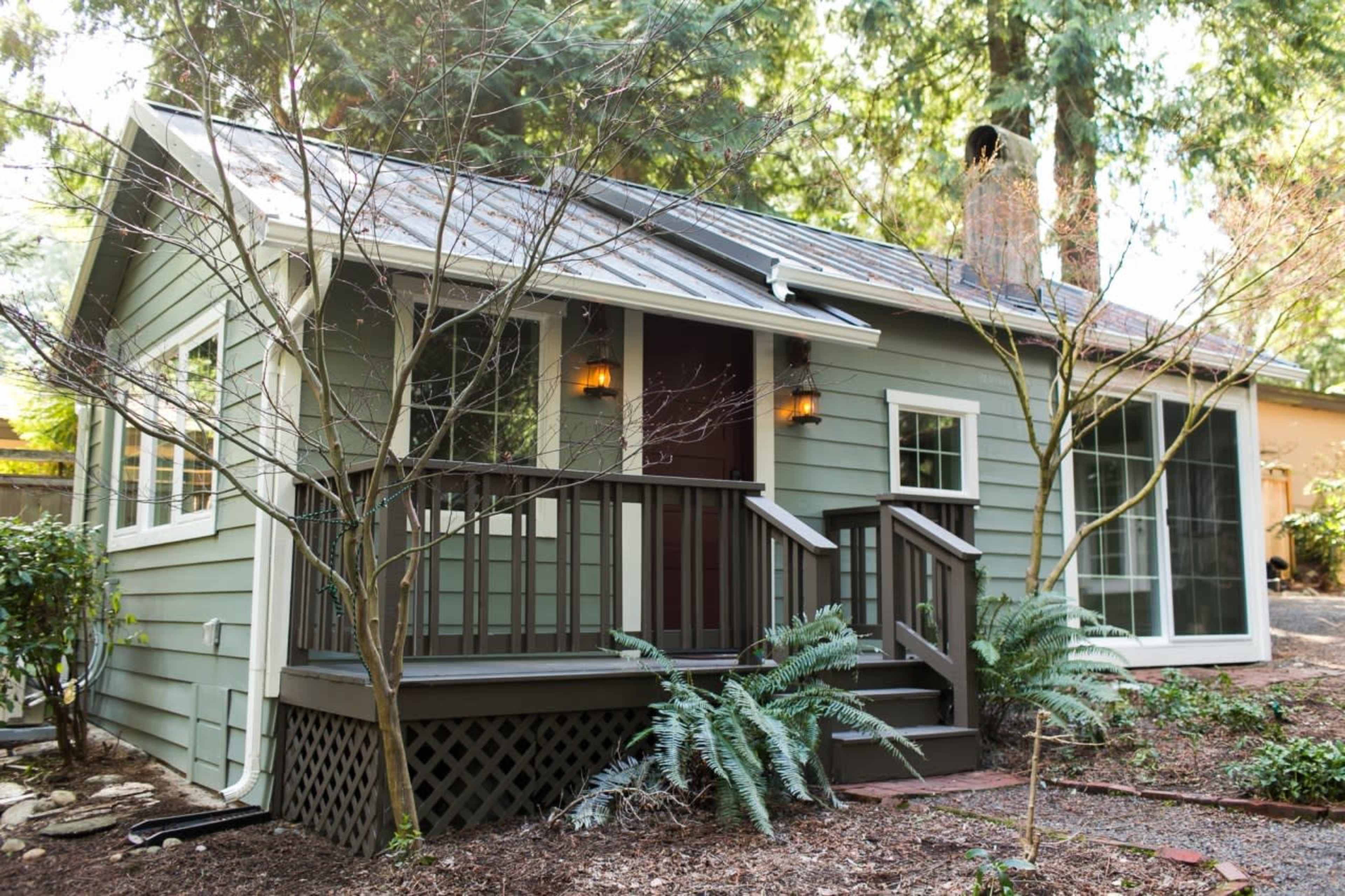 A small green house with a sloped roof and a front porch surrounded by trees and shrubs.