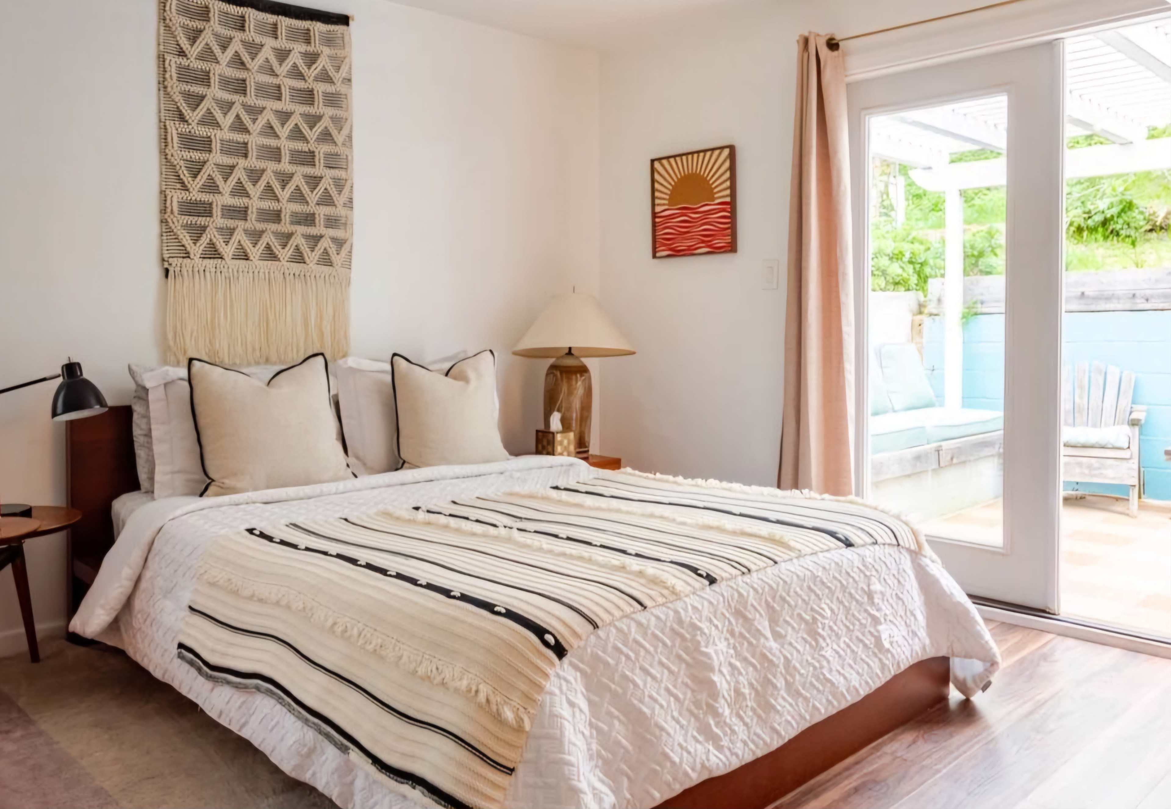 The image shows a cozy bedroom with a bed featuring a patterned blanket, decorative pillows, and a wall hanging above the headboard, along with a light-filled balcony accessible through sliding glass doors.