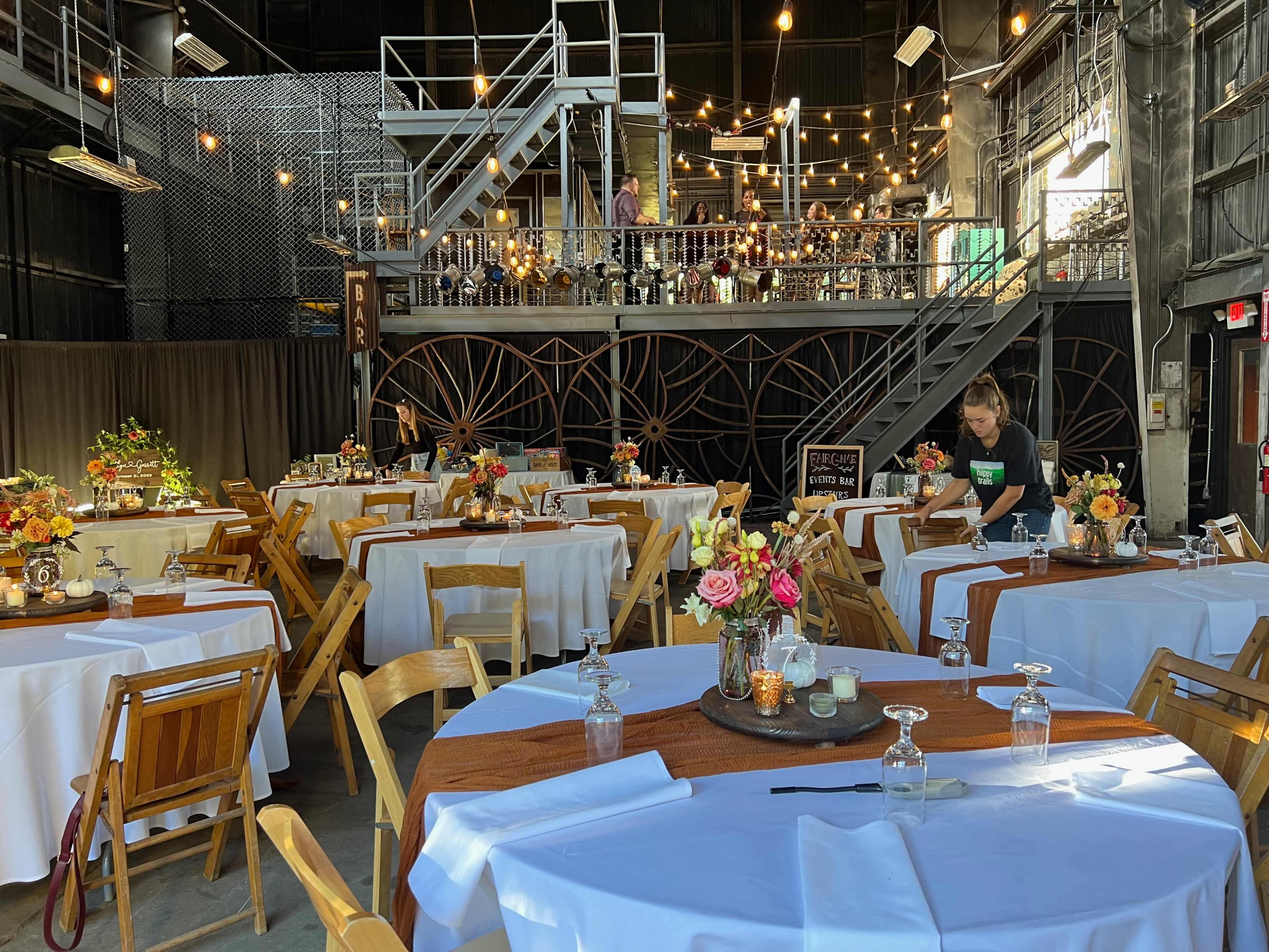 A spacious dining area is set up with tables adorned with white tablecloths and centerpieces, while a server arranges decorations in a venue with exposed wooden features and string lights.