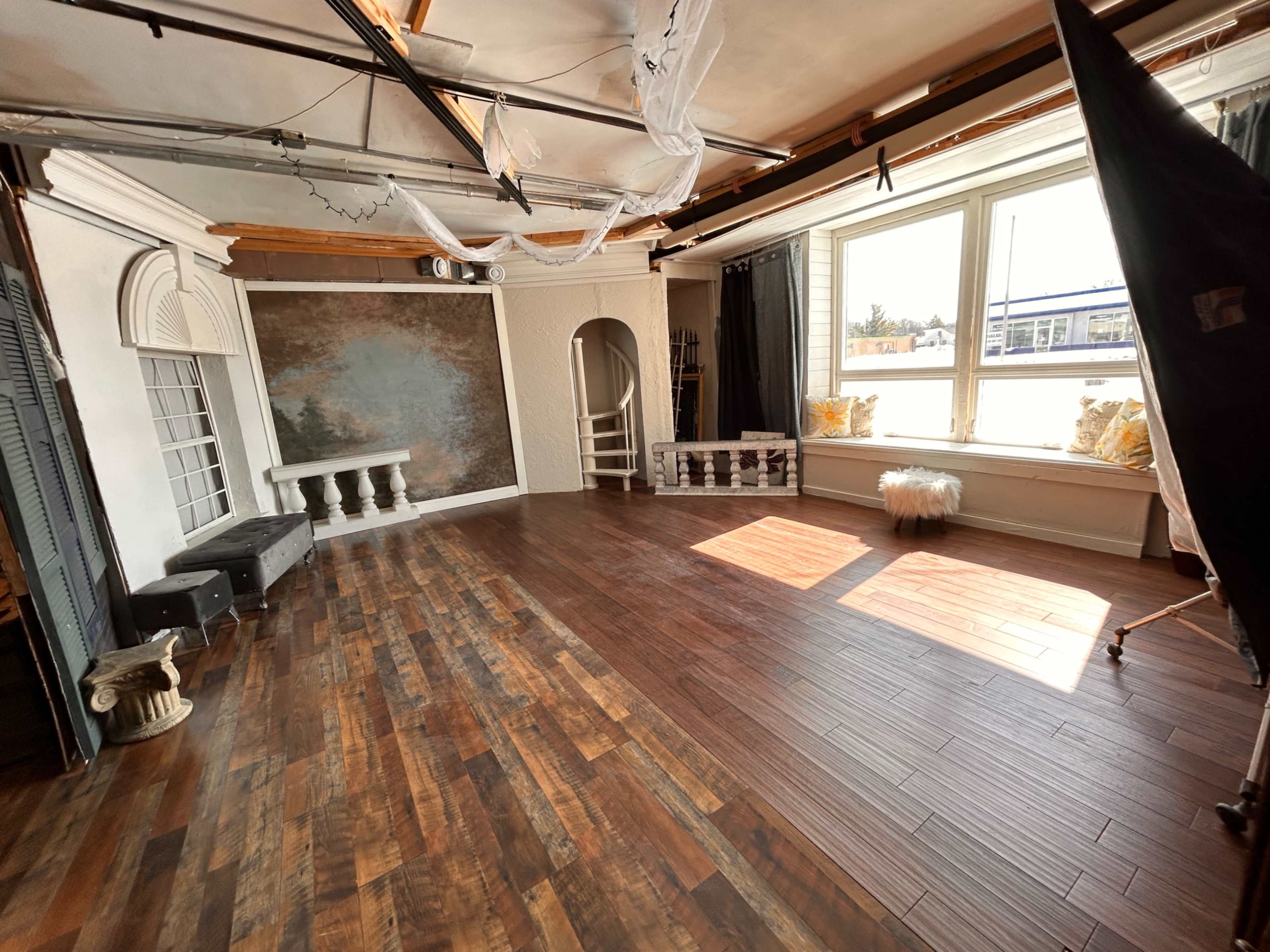 The image shows a spacious photo studio with wooden flooring, a large window, and decorative elements like a stone pedestal and a textured backdrop.