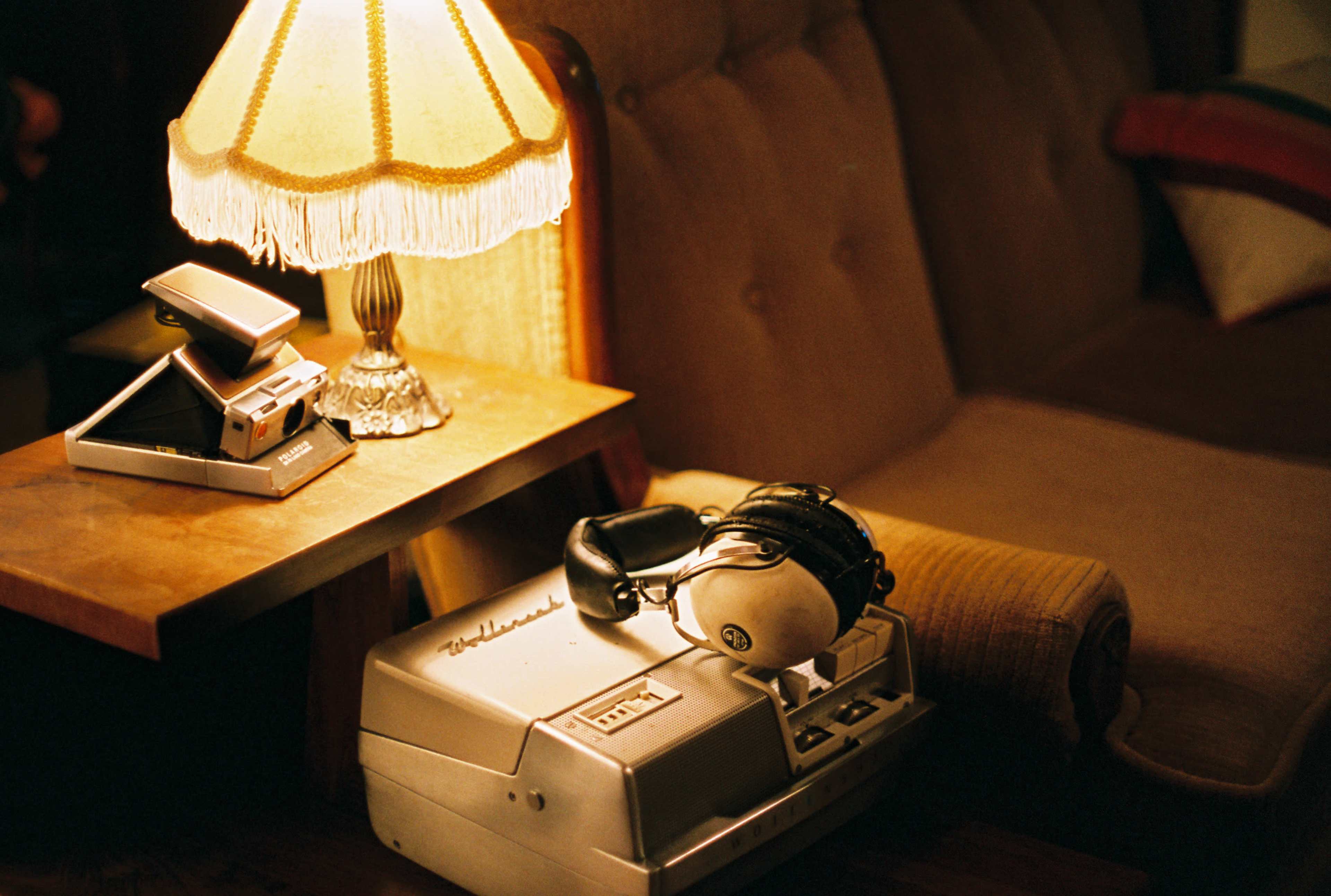 A vintage lamp illuminates a wooden side table with a stapler, headphones, and a reel-to-reel tape recorder next to a couch.