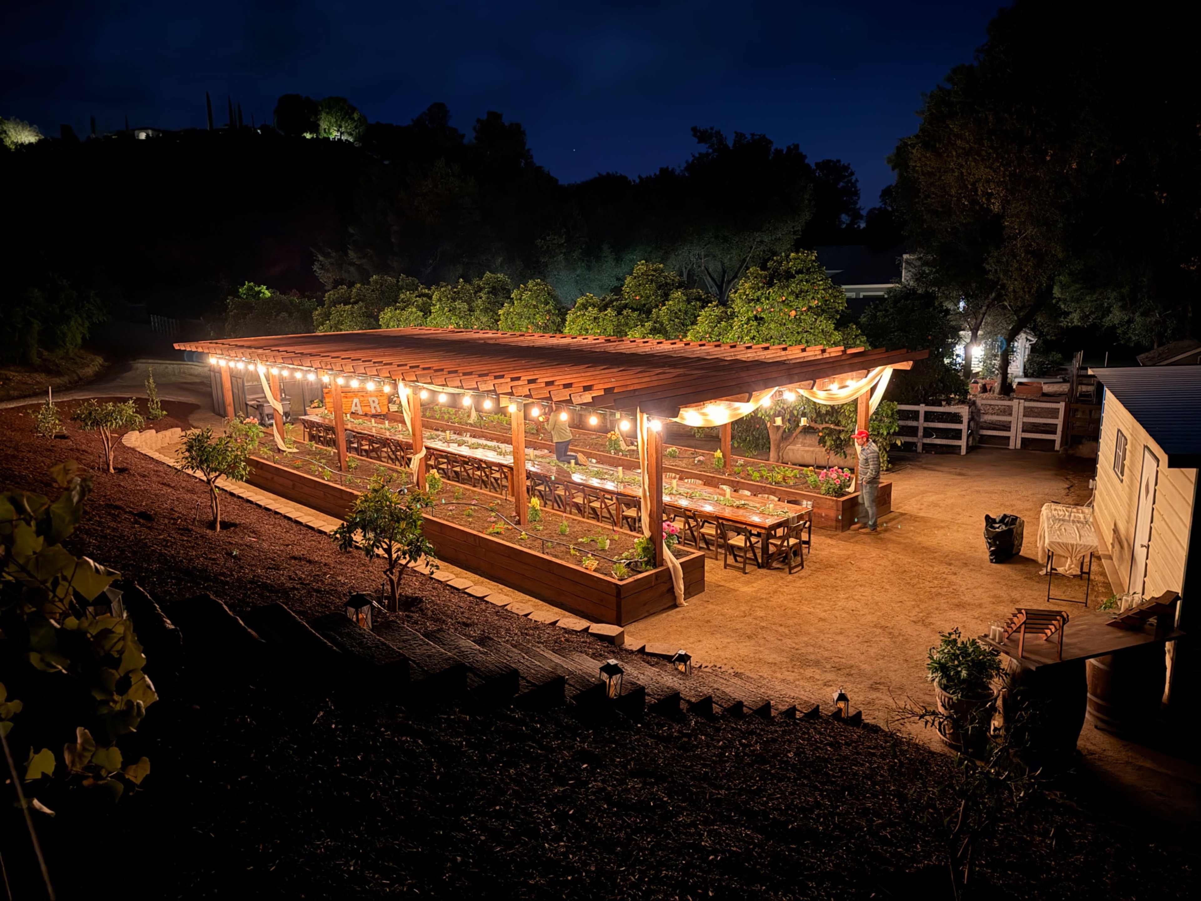 A beautifully lit outdoor dining area with wooden tables and string lights surrounded by lush greenery at night.