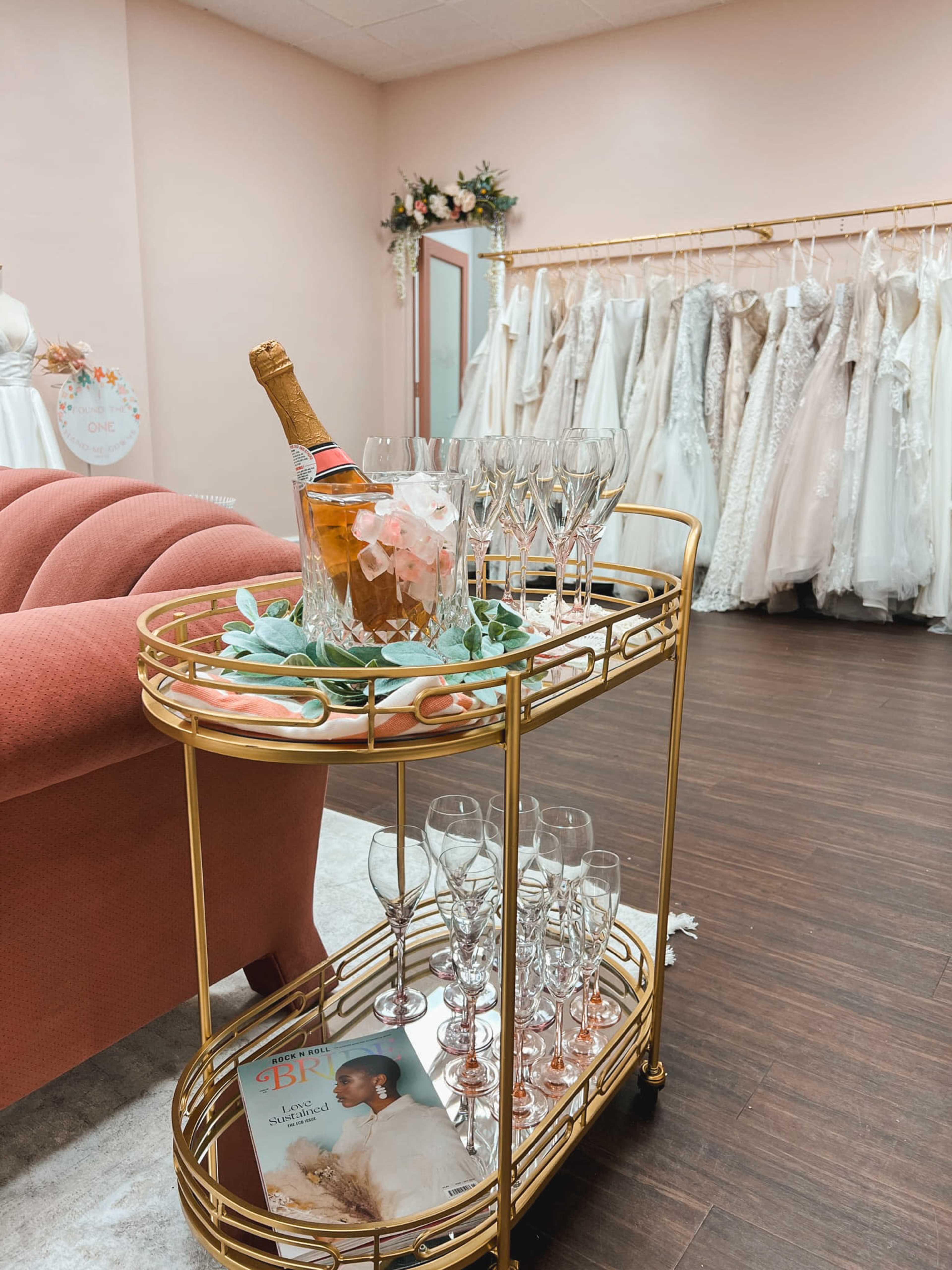 A gold bar cart displays a bucket of champagne surrounded by fluted glasses, with a magazine placed nearby, in a room featuring a rack of wedding dresses.