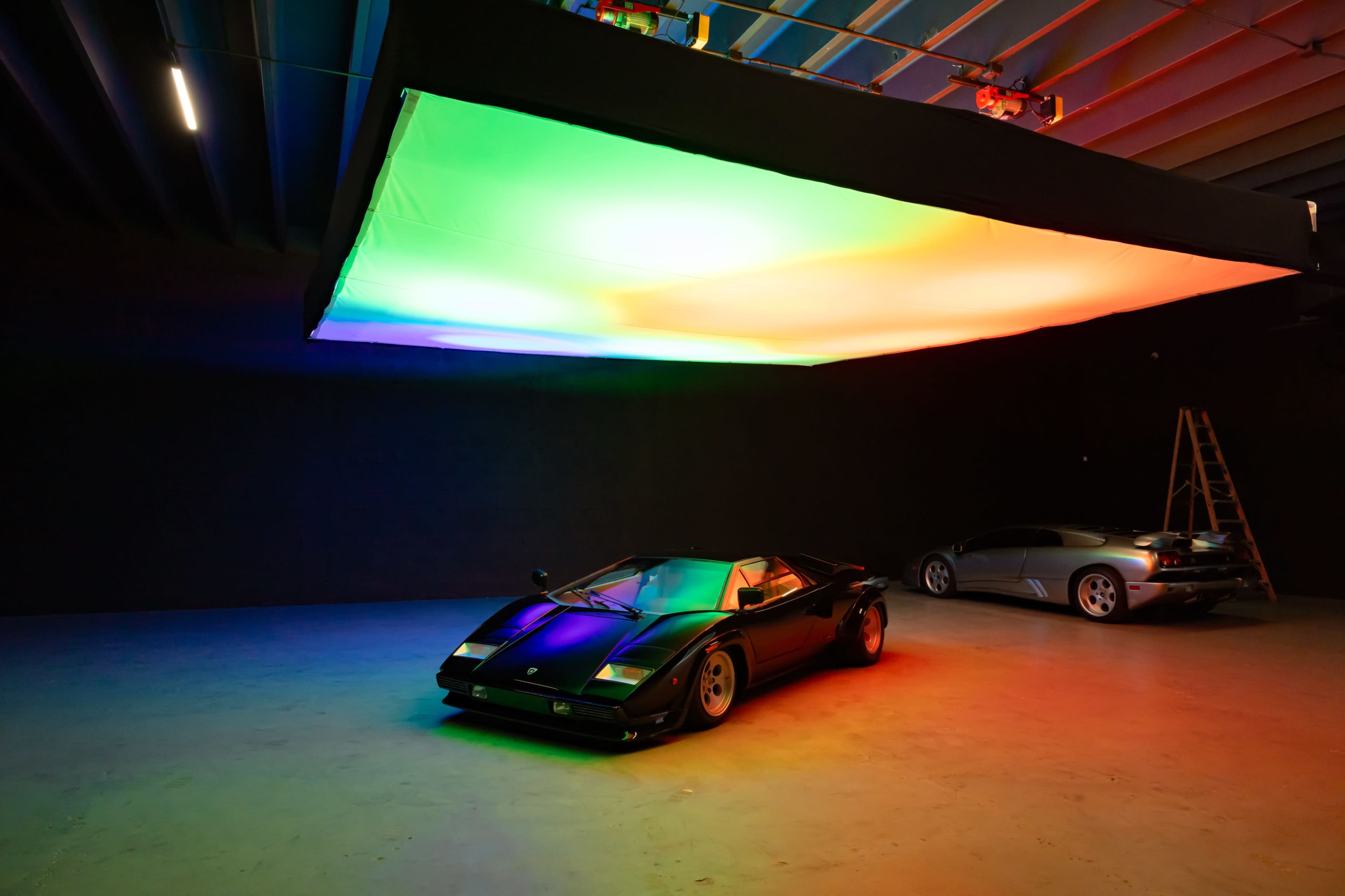 A black sports car is positioned under a colorful illuminated ceiling in a dimly lit studio, with a silver car visible in the background.