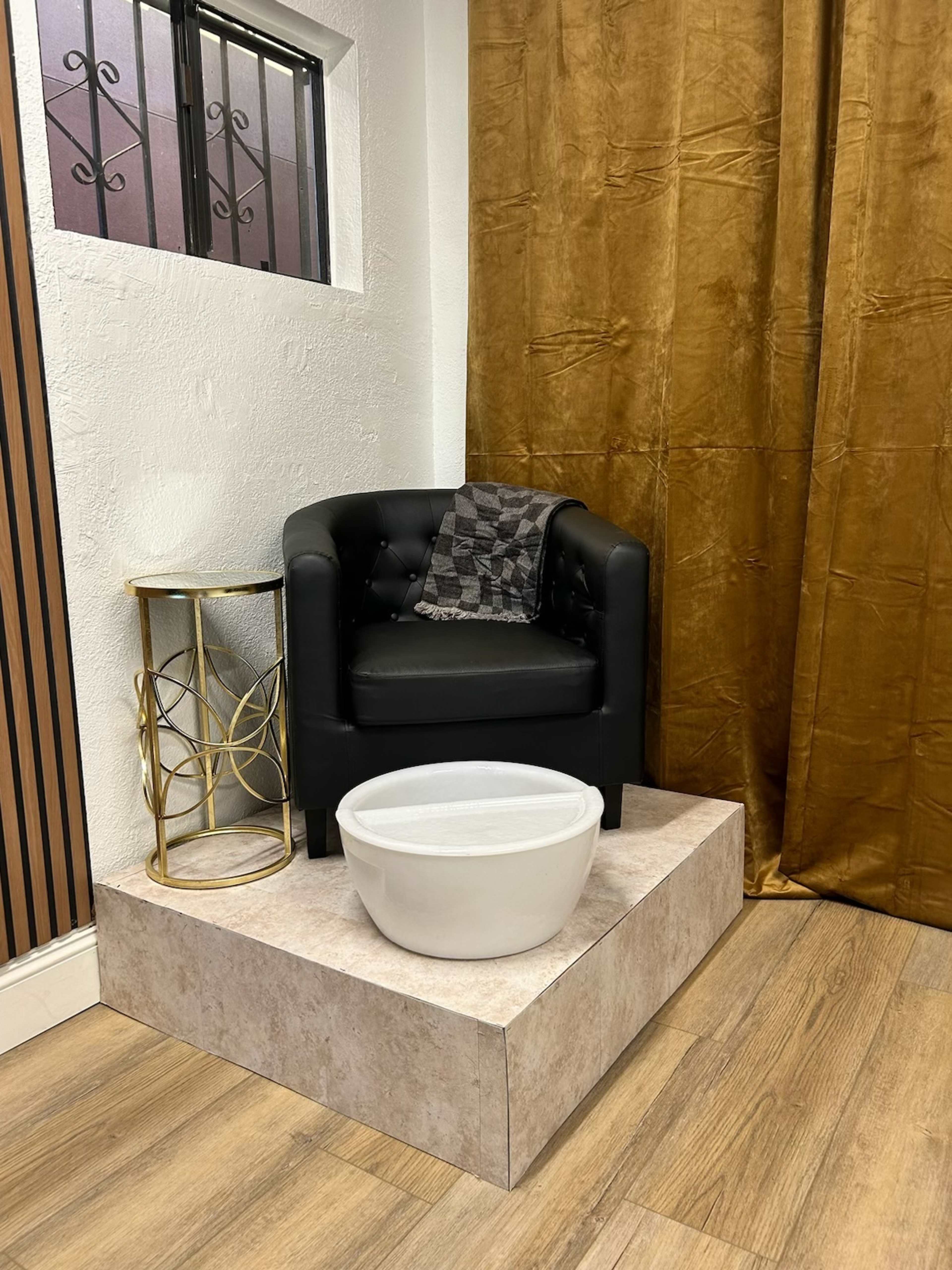 A black upholstered chair is positioned beside a circular decorative table and a white basin on a raised platform in a room with a gold curtain.