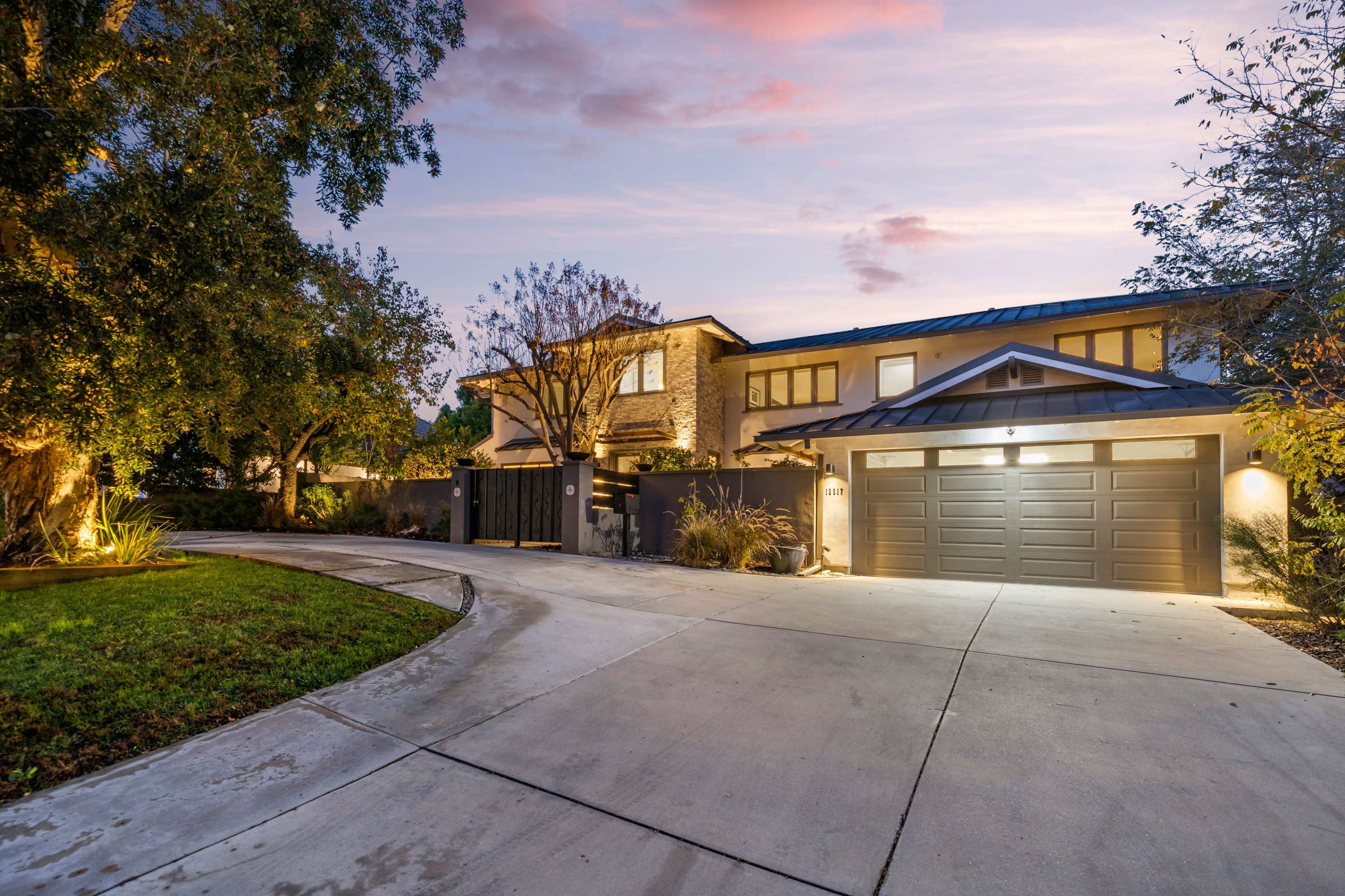 A modern two-story house with a gray garage, surrounded by trees and a circular driveway at dusk.