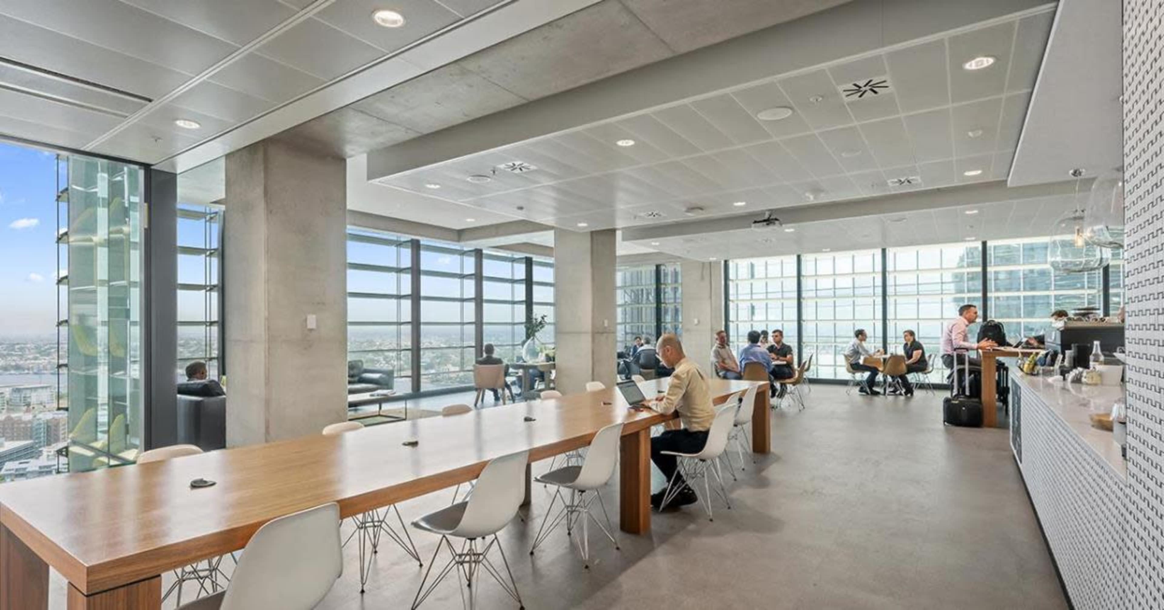 A modern office space features a long table with chairs, large windows showcasing a city view, and several workers engaged in various activities.
