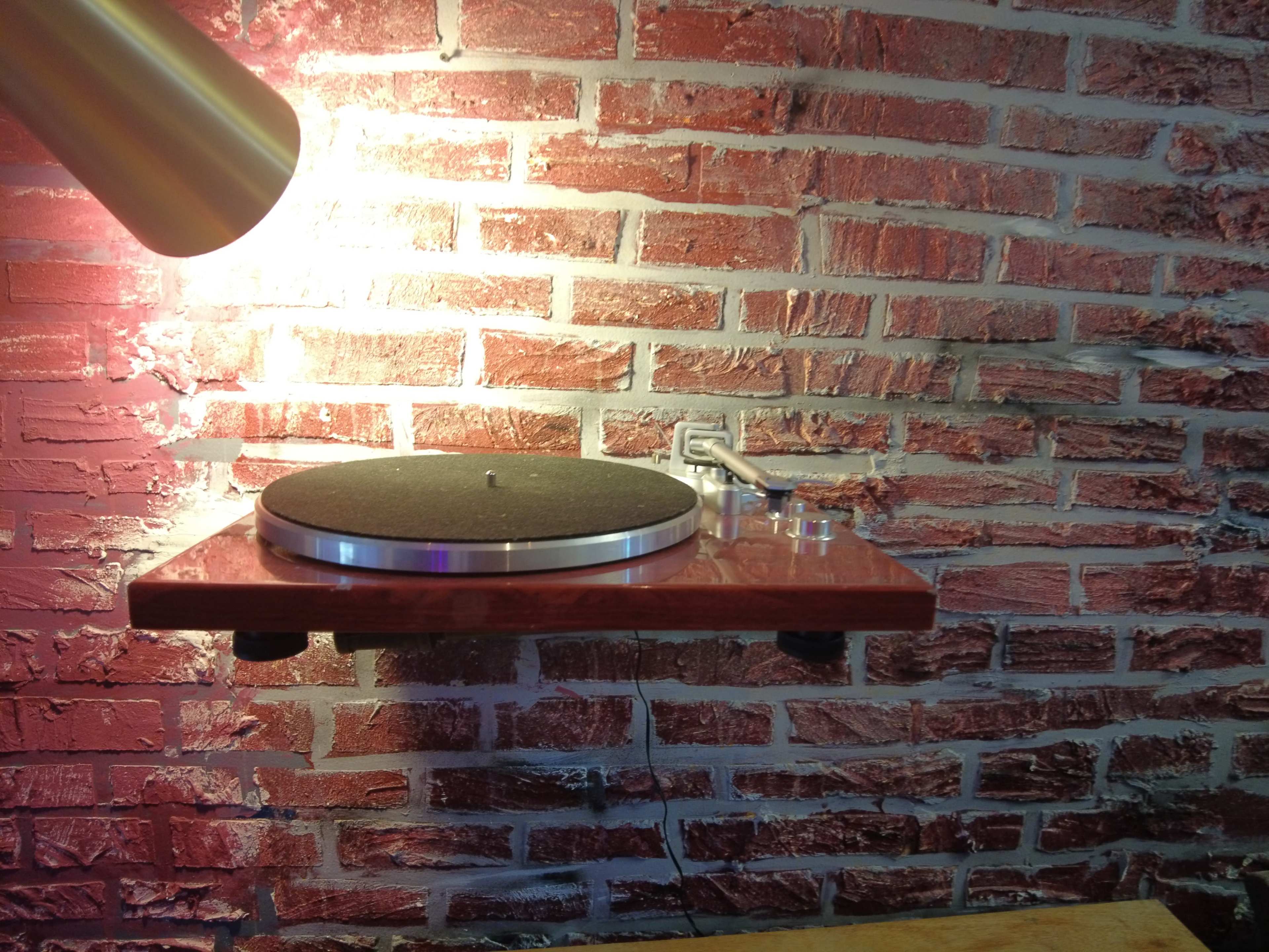A turntable is mounted on a wooden shelf against a brick wall, illuminated by a nearby lamp.
