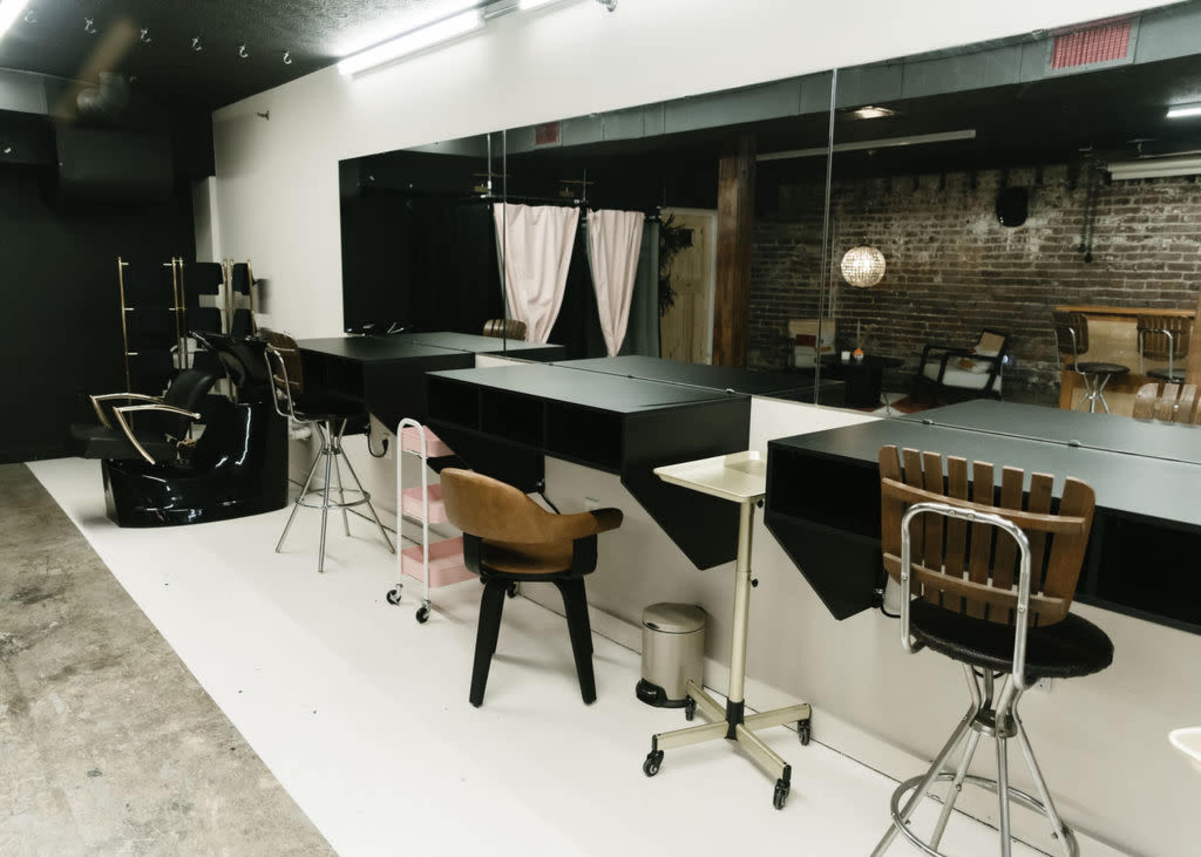 The image shows a modern hair salon interior featuring multiple styling stations with chairs, large mirrors, and a mix of black and wooden furniture against a brick wall.