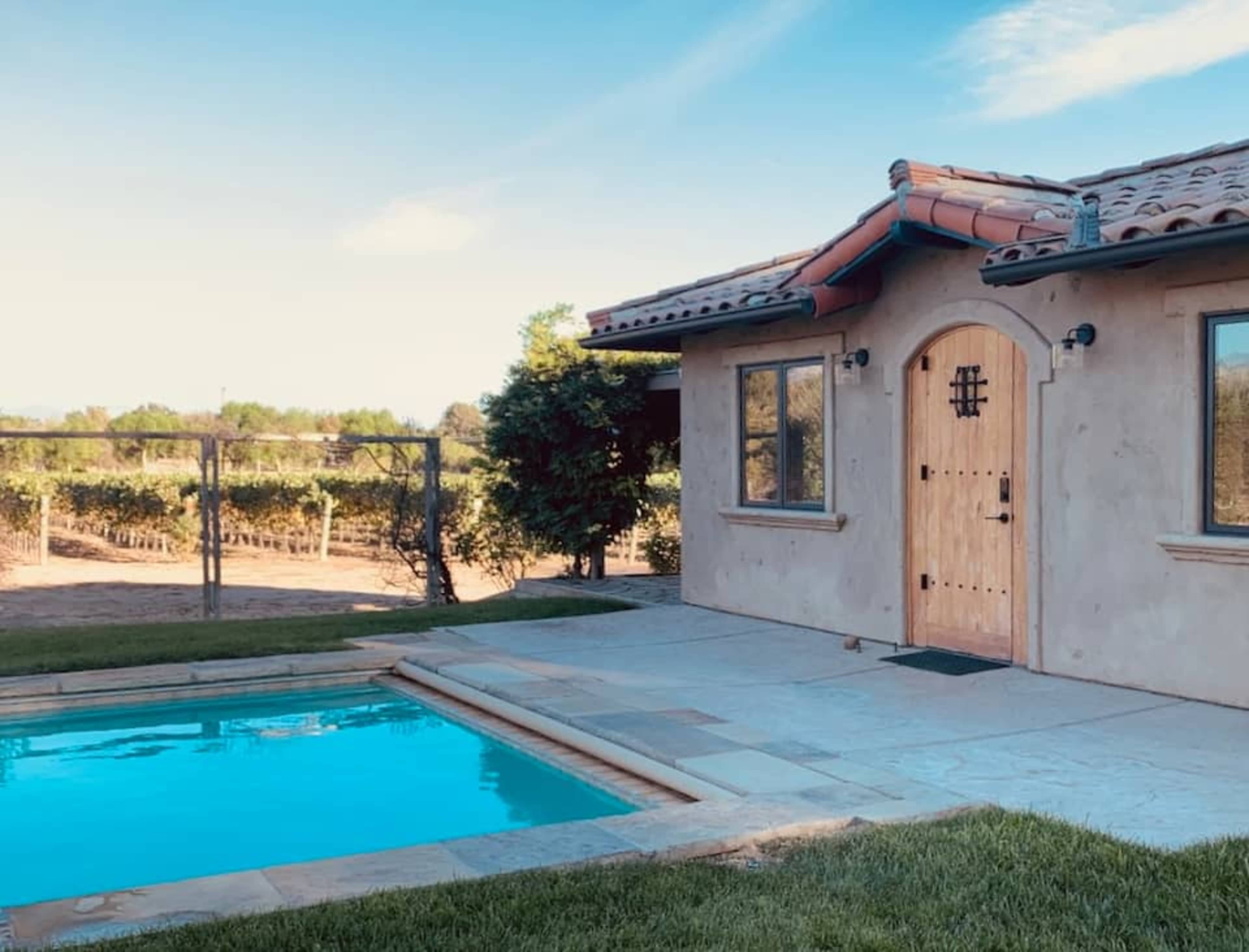 A house with a tiled roof beside a swimming pool, surrounded by a grassy area and vineyards in the background.