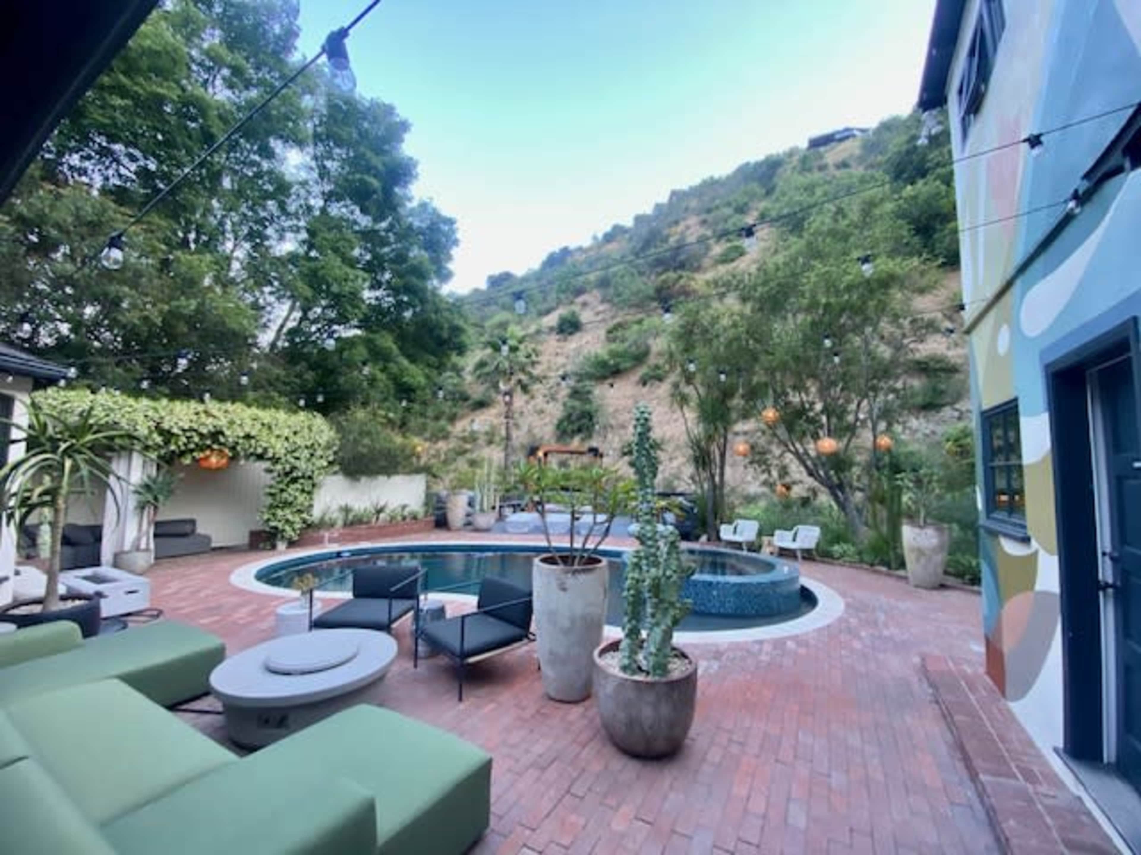 The image shows a backyard with a circular pool surrounded by lounge chairs and potted plants, nestled against a hillside.
