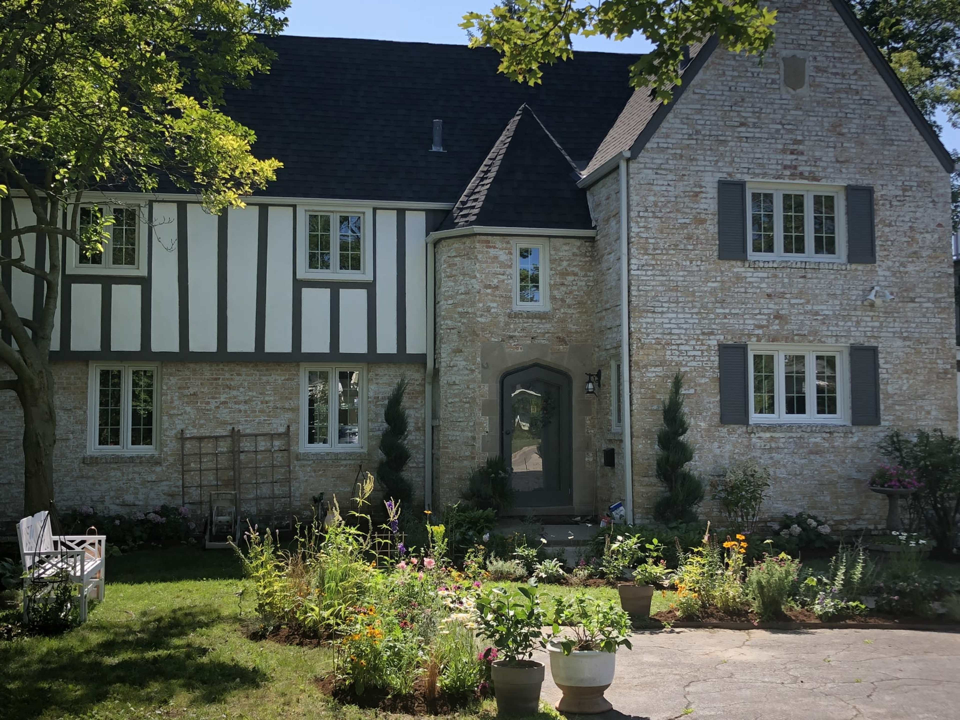 A two-story house with a mix of stone and stucco exterior, surrounded by a garden filled with various plants and flowers.