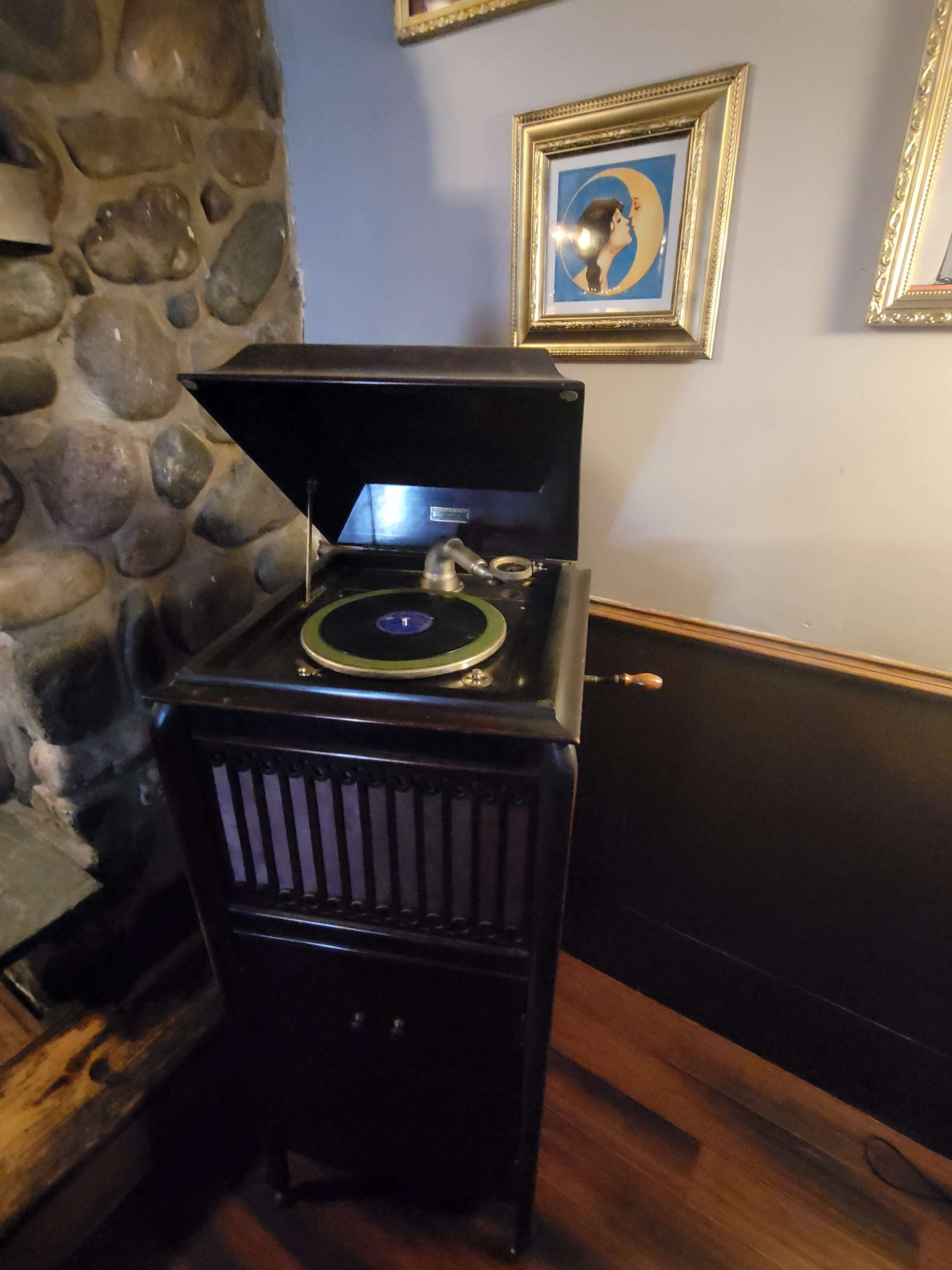 A vintage phonograph with a closed lid is positioned next to a stone wall, under a framed portrait on the wall.