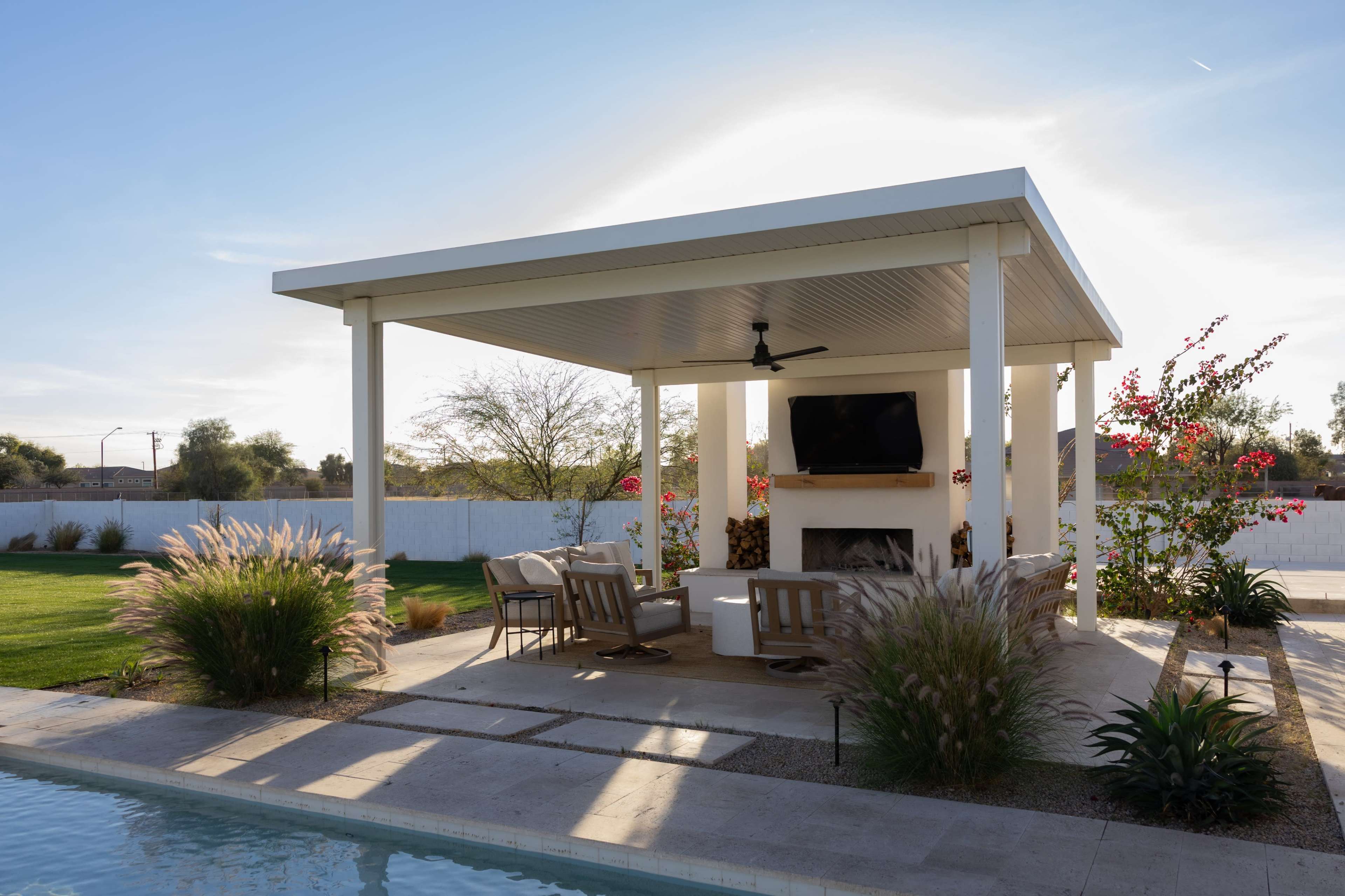 A modern outdoor gazebo with a ceiling fan and a television, surrounded by landscaped greenery and a pool.