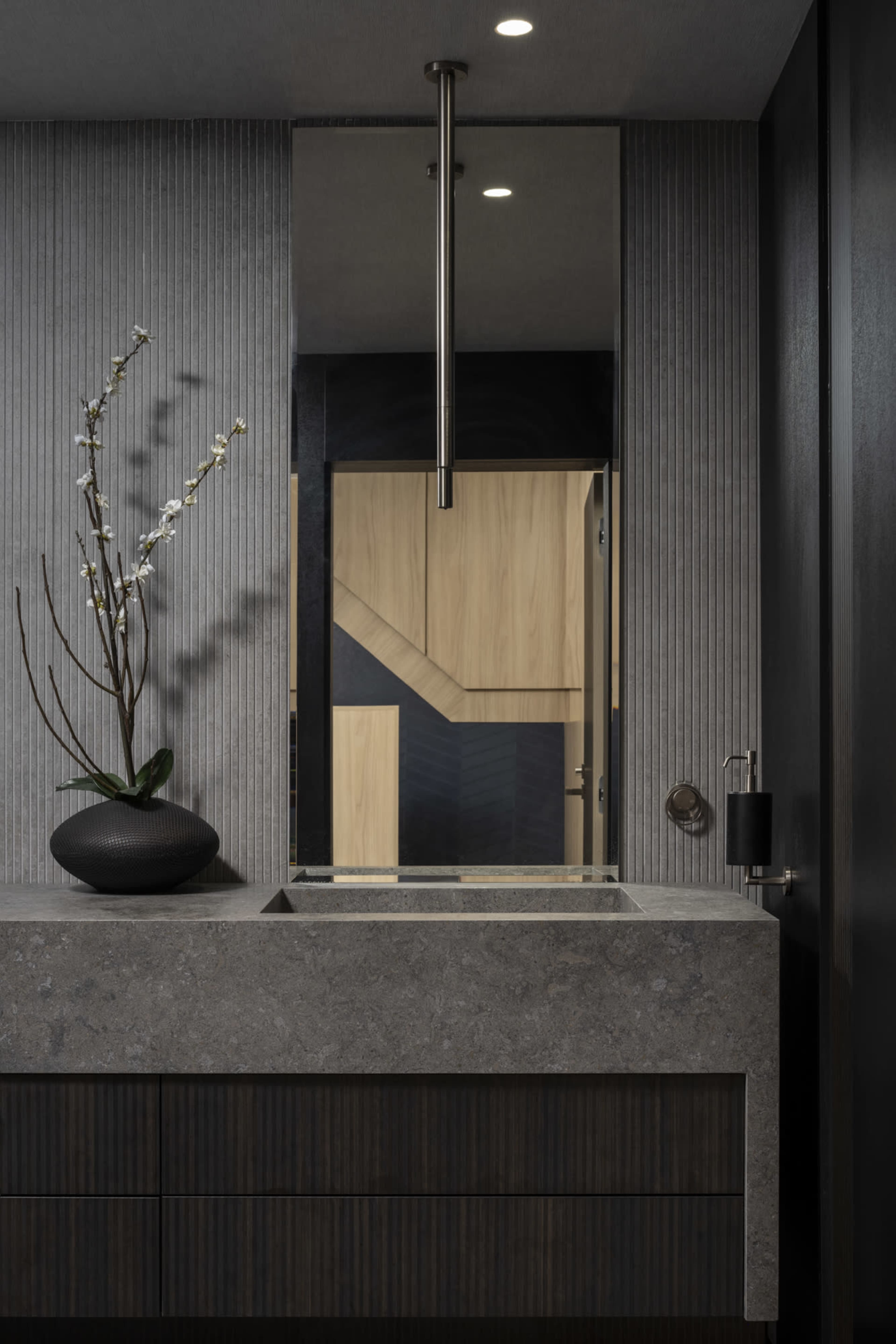 The image shows a modern bathroom featuring a sleek, stone countertop with a dark vase containing a flower, a large mirror hanging above, and a subtle, striped wall design.