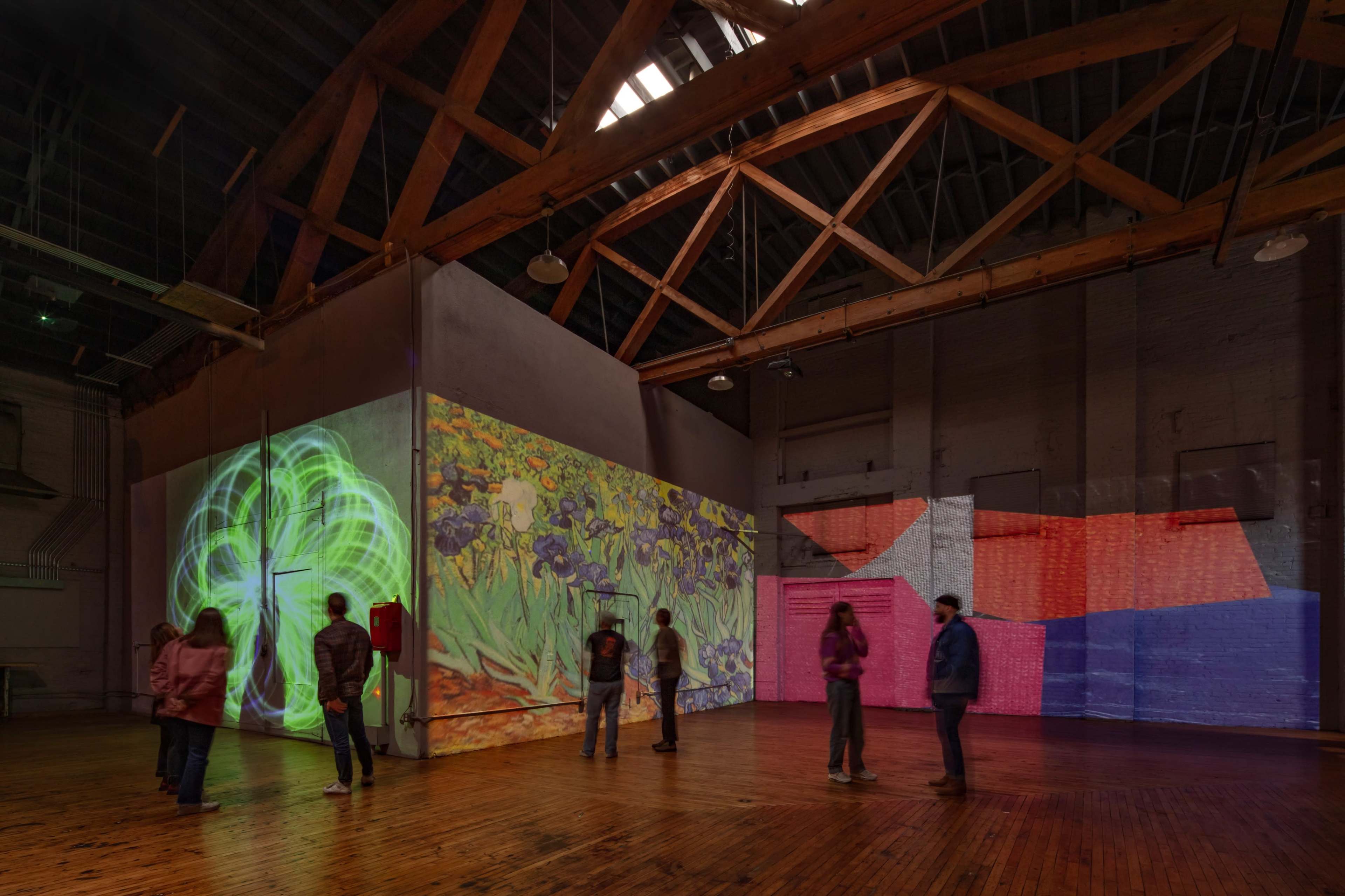 A large, open industrial space features colorful projections on the walls, with several people observing the displays.