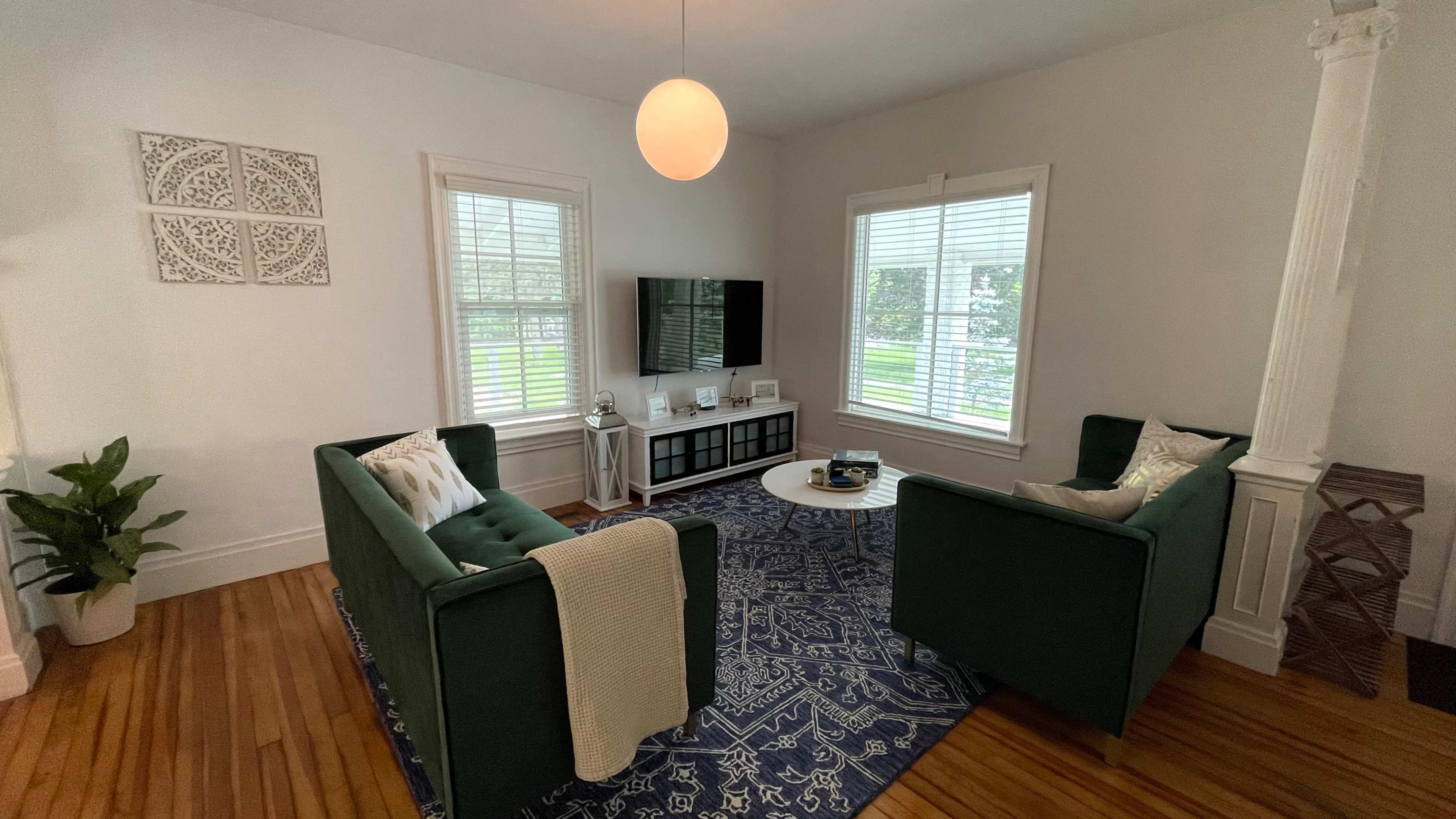 A cozy living room with two green sofas facing a coffee table, a television mounted on the wall, and large windows letting in natural light.